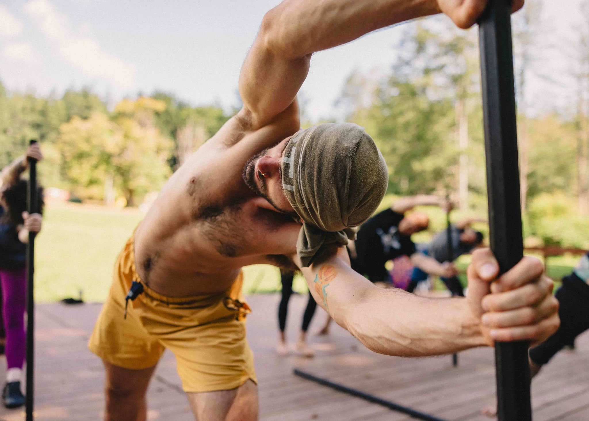 A shirtless man with a bandana performs a yoga pose outdoors, holding onto black poles with trees in the background.