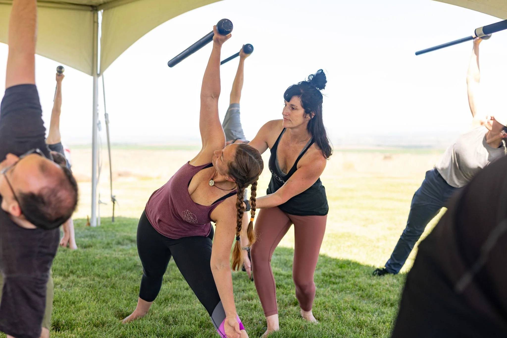 Women participating in an outdoor yoga or fitness class under a canopy, with some performing poses using small black weights.