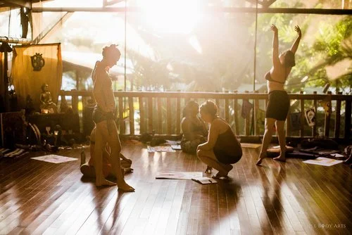 Three women practicing yoga or dance in a studio with sunlight streaming through large windows, wooden floors, and various equipment and decorations visible.