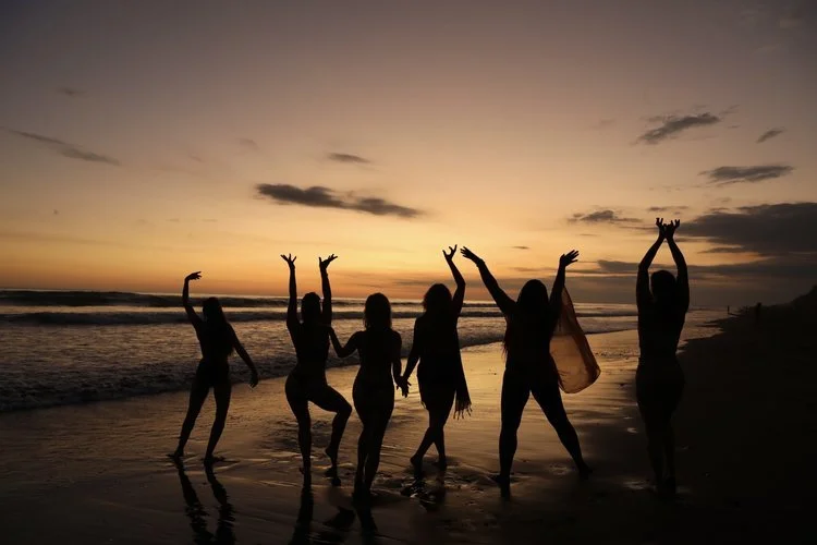 Group of six women dancing and celebrating on the beach at sunset.