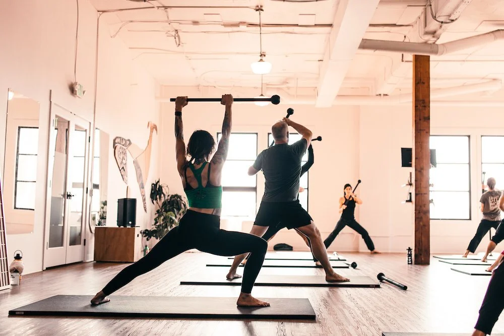 People participating in a group yoga class with some holding workout equipment in a spacious, well-lit studio.