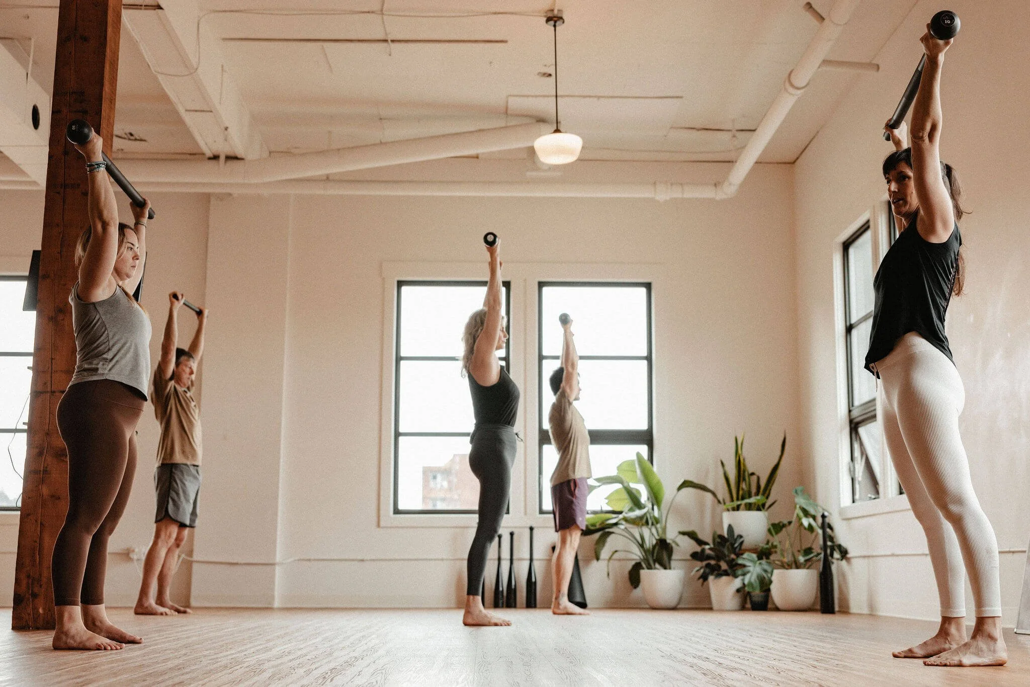 Group of people participating in a yoga or fitness class, raising small dumbbells overhead in a spacious studio with large windows and indoor plants.