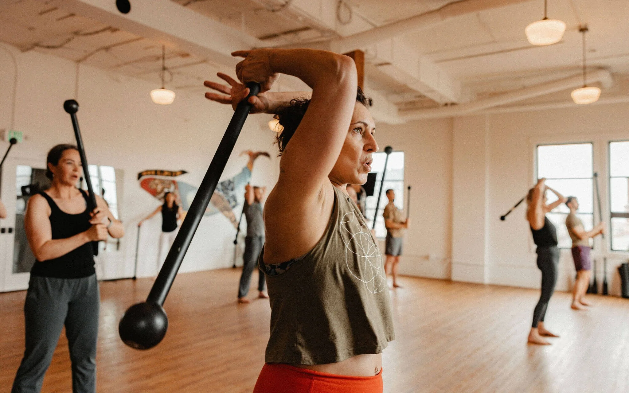 A group of people participating in a fitness class indoors, with a woman in the foreground holding a black exercise stick overhead.