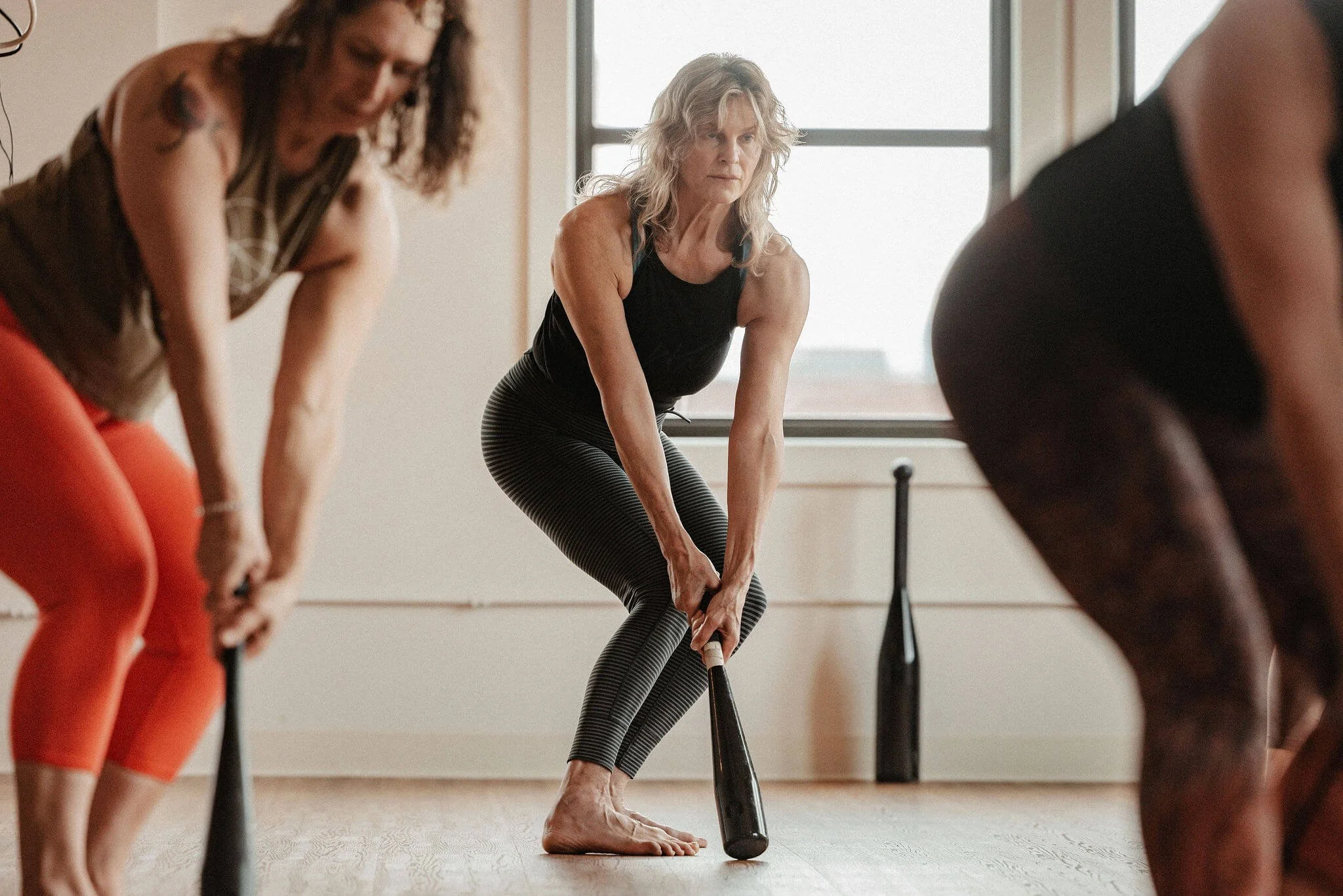Women participating in a yoga class using paddles, seen on a wooden floor with large windows in the background.