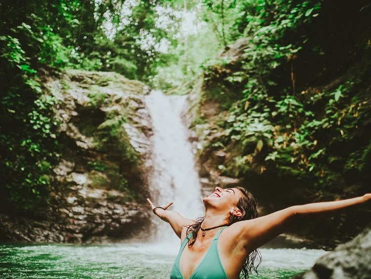 A woman with arms outstretched enjoying a waterfall surrounded by lush green trees in a forest.