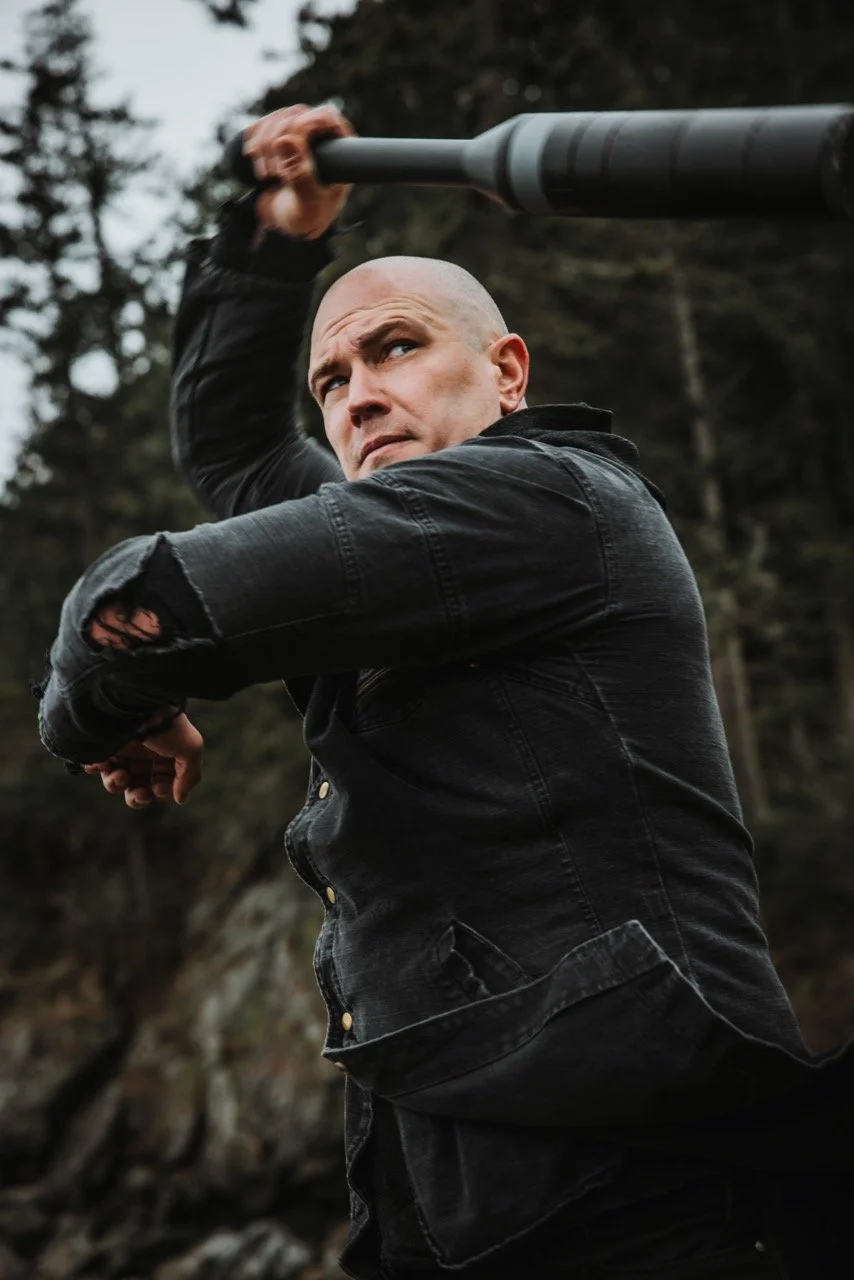 A bald man in a black, ripped jacket holding a large baseball bat or club over his shoulder outdoors with trees and rocks in the background.