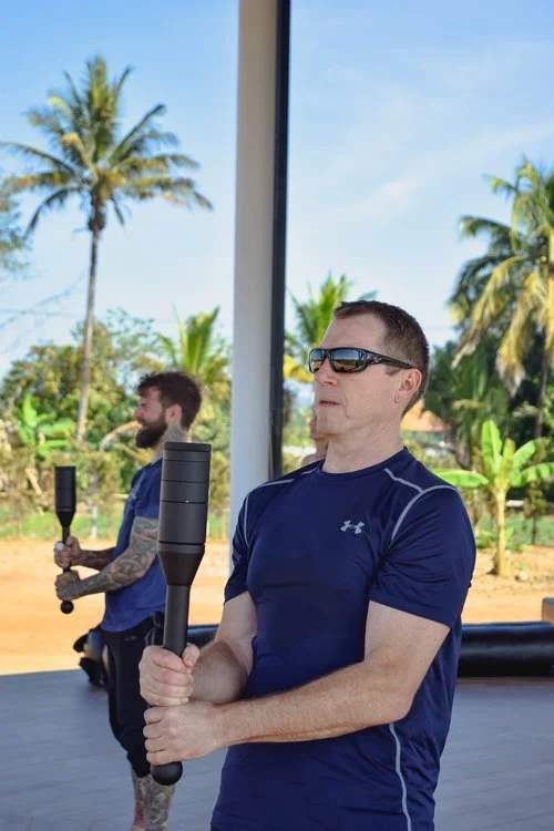 Two men holding black weighted batons indoors, with a window showing trees and a blue sky outside.