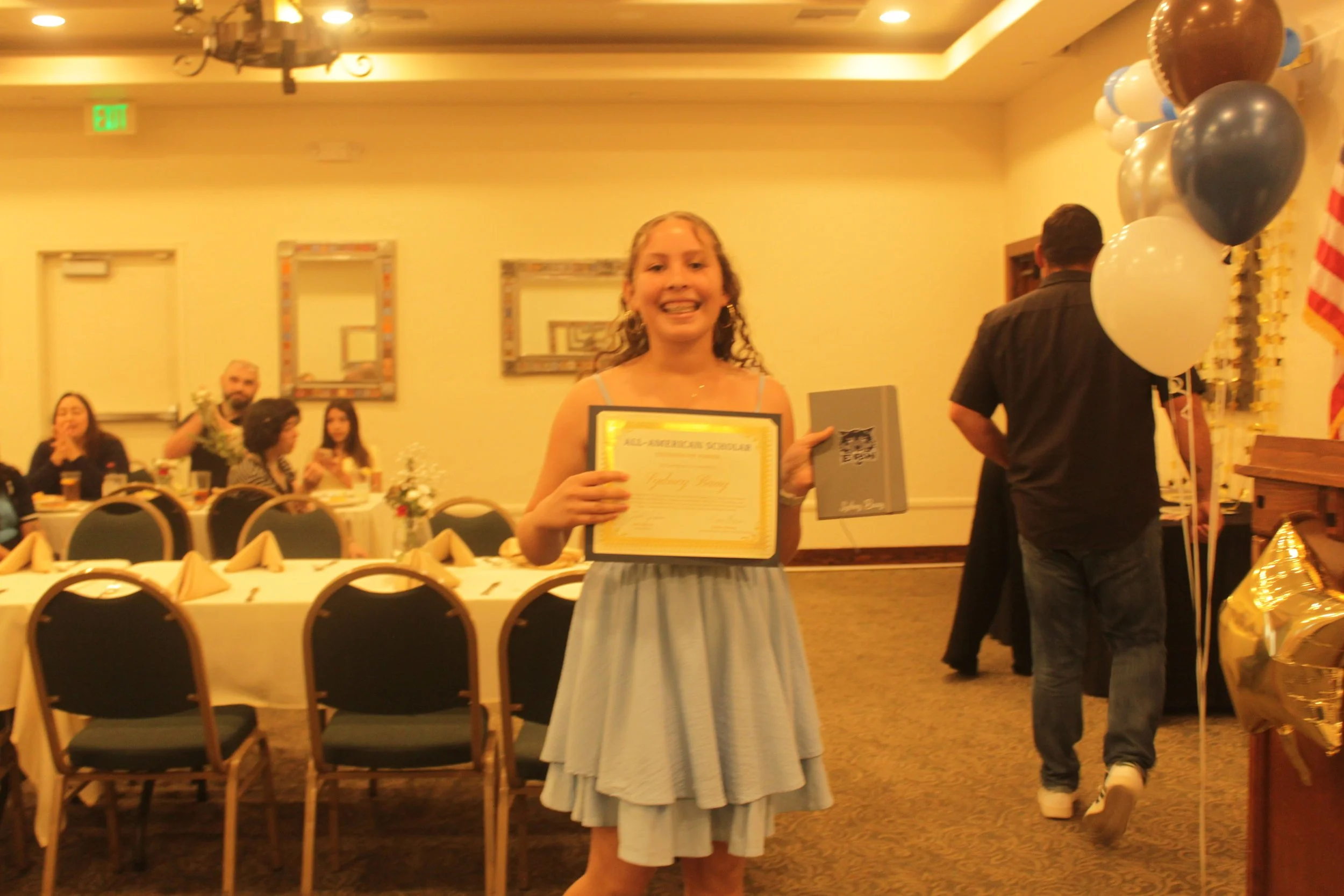 A young girl in a gray dress holding a framed certificate and a small gift box, smiling at a celebrating event in a decorated hall with balloons and people sitting at tables.