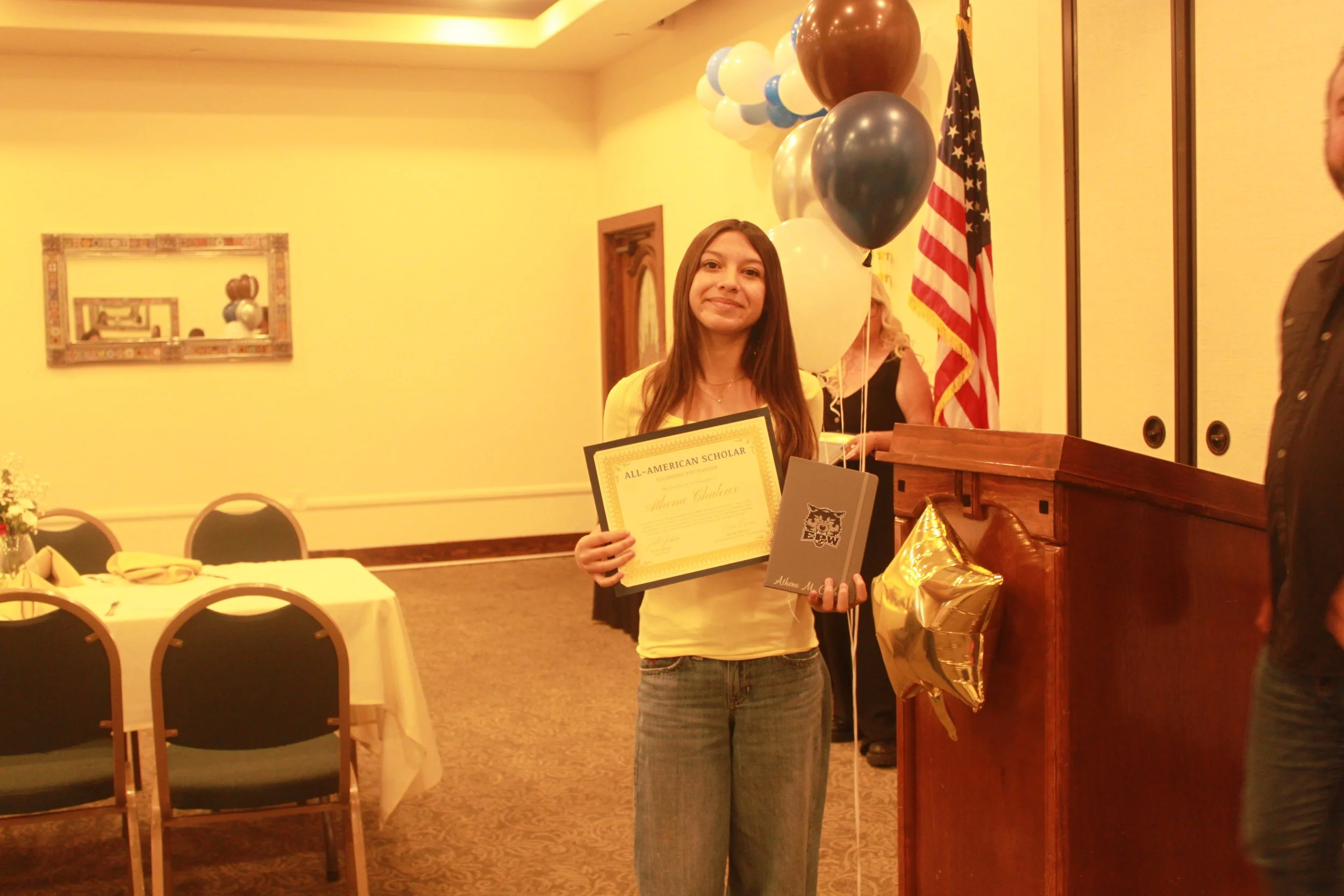 A young girl holding an all-American scholar certificate and a folder, standing in a decorated banquet room with balloons and an American flag.