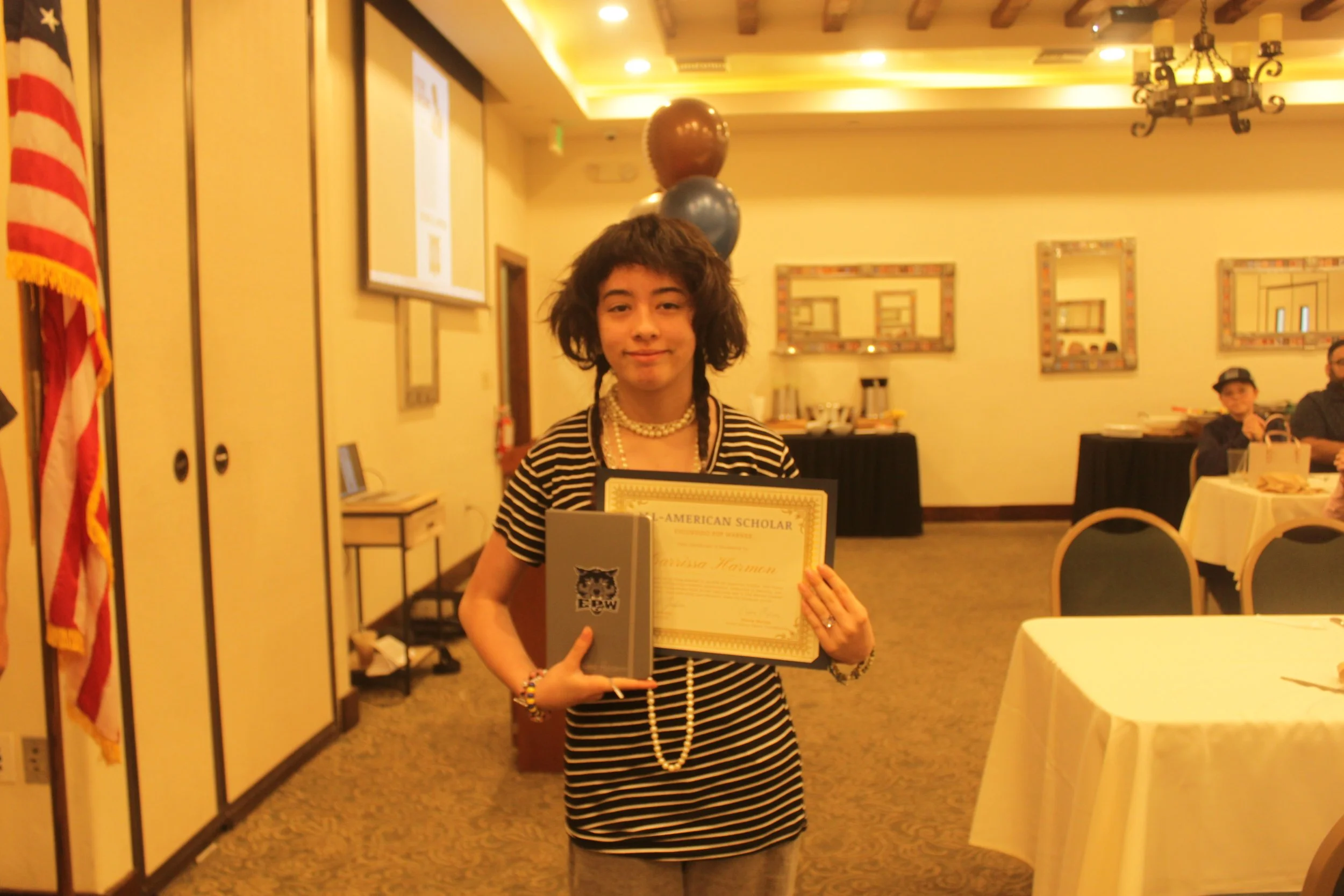 A young person with medium-length curly hair, wearing a striped shirt, multiple pearl necklaces, and bracelets, standing in a banquet room holding a certificate. The background includes banquet tables, a chandelier, balloons, and a small group of people seated at tables.