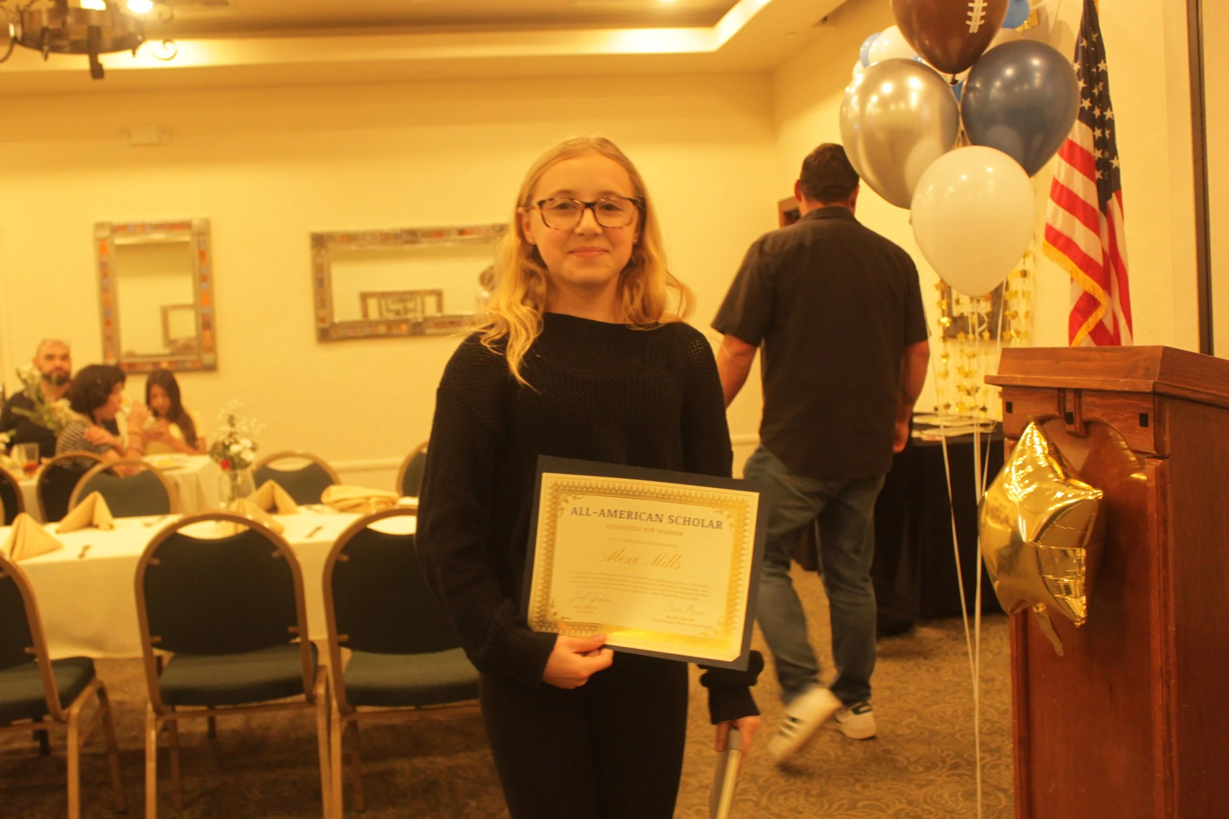 Young woman holding an 'All-American Scholar' certificate at an indoor awards or recognition event with balloons and American flags in the background.