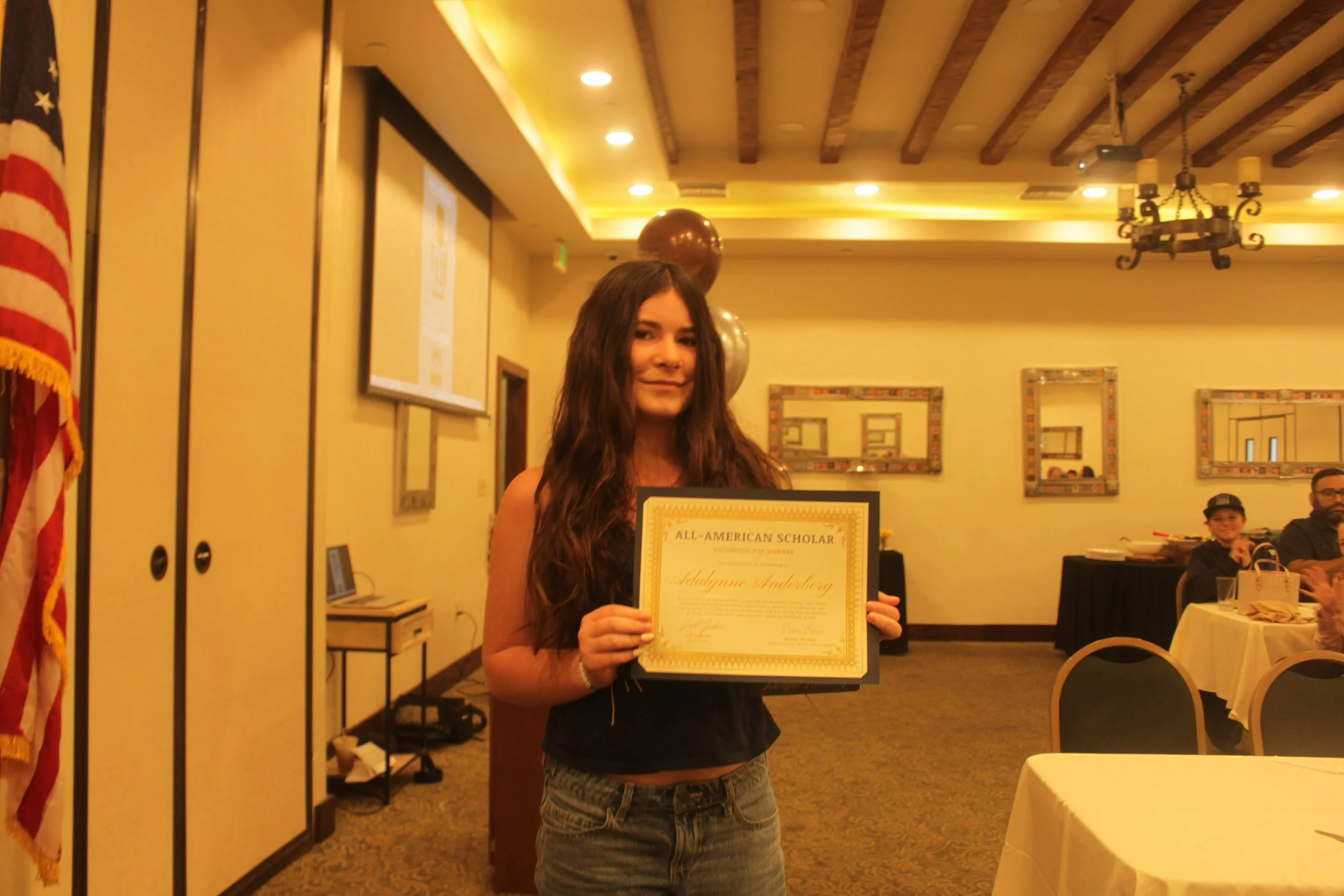 A young woman with long brown hair holding a framed certificate at an awards ceremony in a banquet hall. People are seated at round tables in the background, and decorative mirrors hang on the wall.