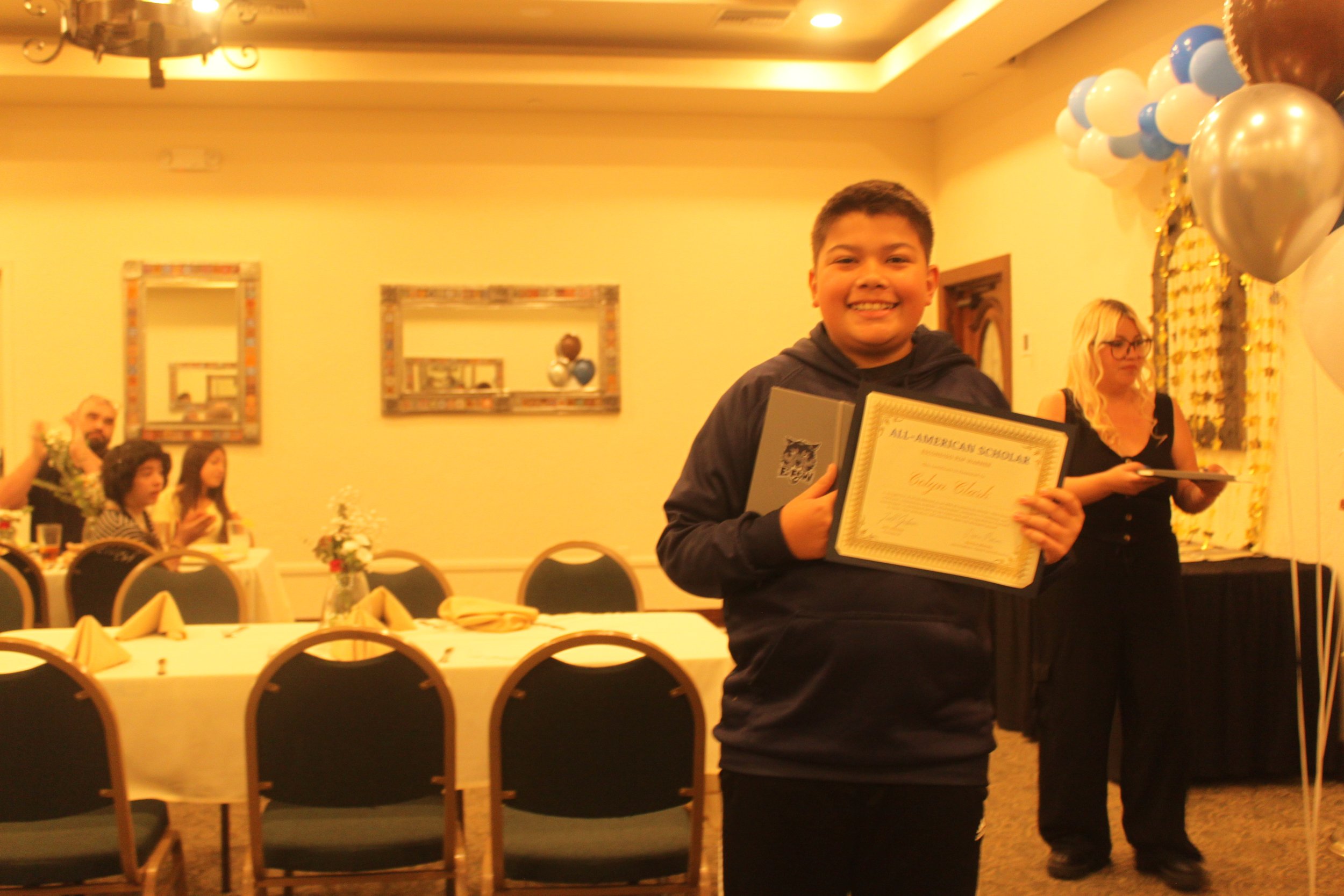 A young boy holding a certificate at an indoor celebration, with balloons and people in the background.