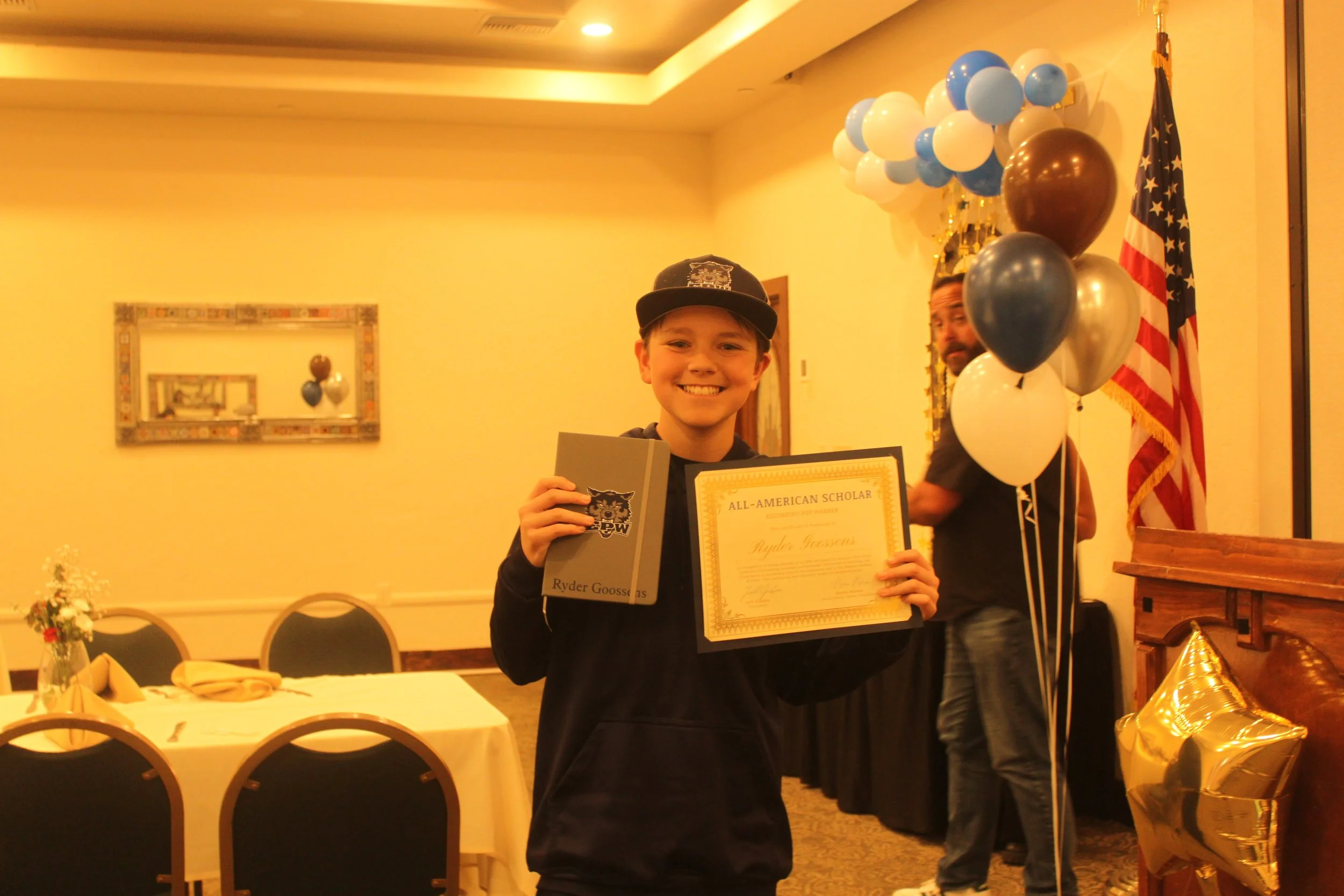 A young boy smiling and holding a certificate and a folder at an awards ceremony. There are balloons and an American flag in the background.