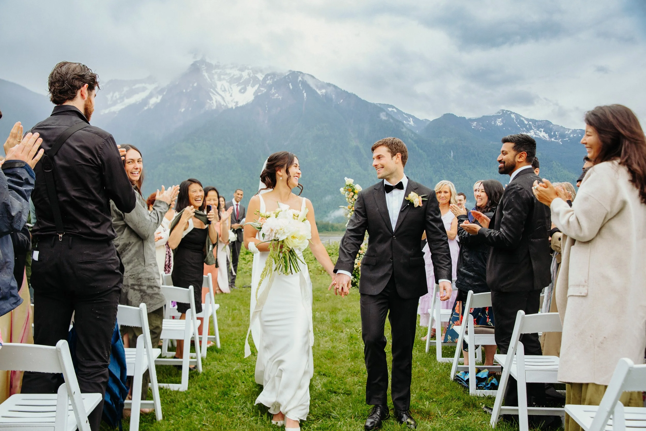 Wedding ceremony outdoors with mountains in the background. Bride and groom holding hands, surrounded by guests clapping and smiling.