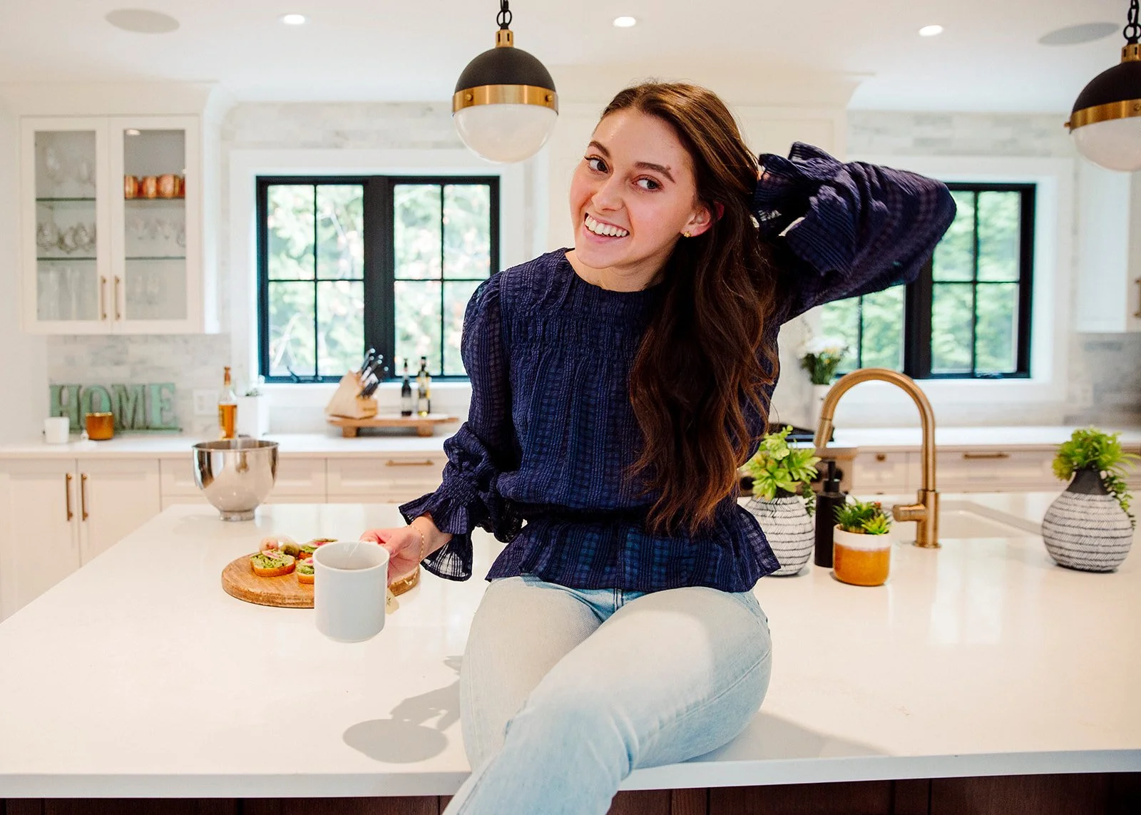 A young woman with long brown hair and a smile is sitting on a kitchen island, holding a white mug in her right hand, with a plate of snacks and two glass jars nearby. The kitchen has white cabinets, a window with black frames, and potted plants on t