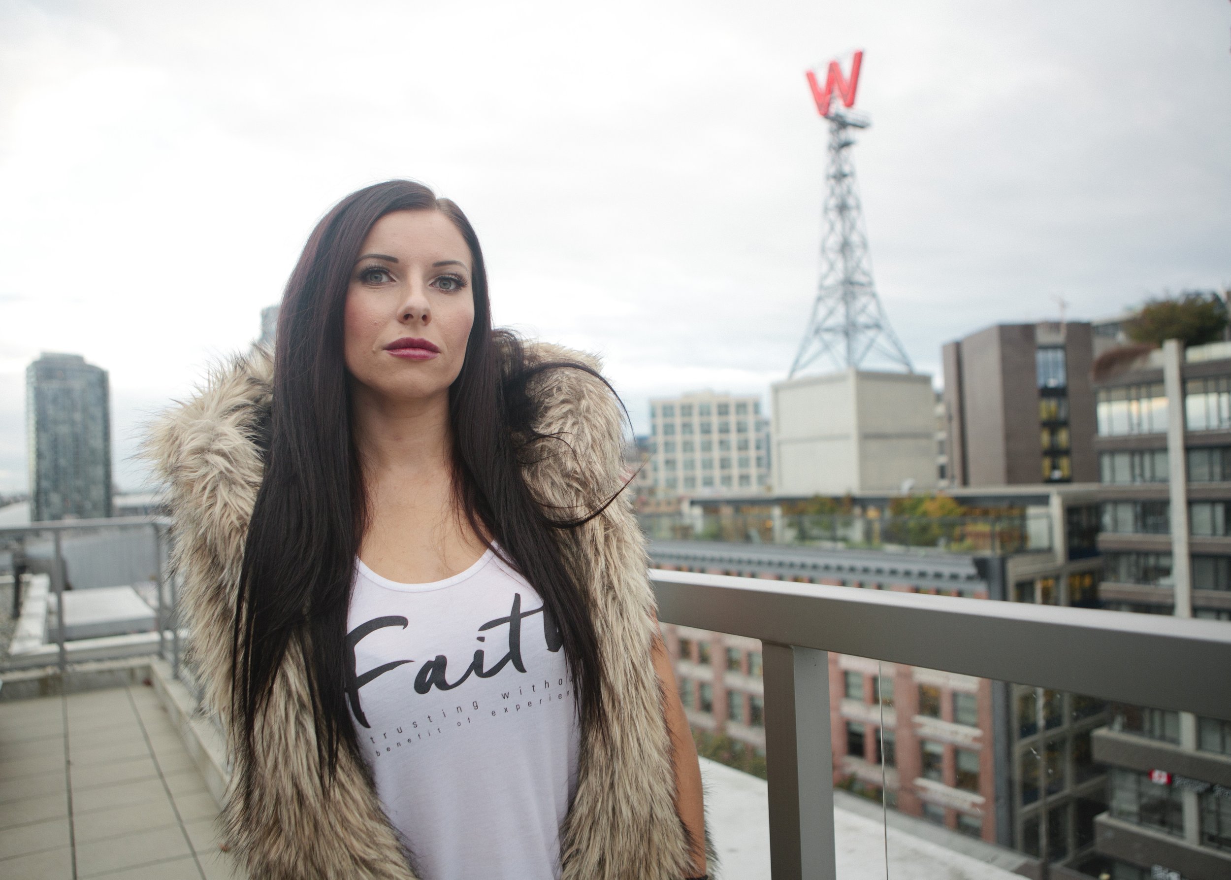 A woman with long dark hair and light skin standing on a rooftop terrace with a cityscape background, wearing a white T-shirt with the word 'Faith' written on it and a fur-lined jacket.