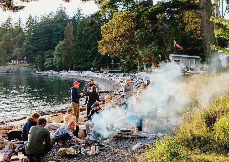 People gathered around a campfire on a rocky lakeshore, with trees and a small house in the background, during sunset.