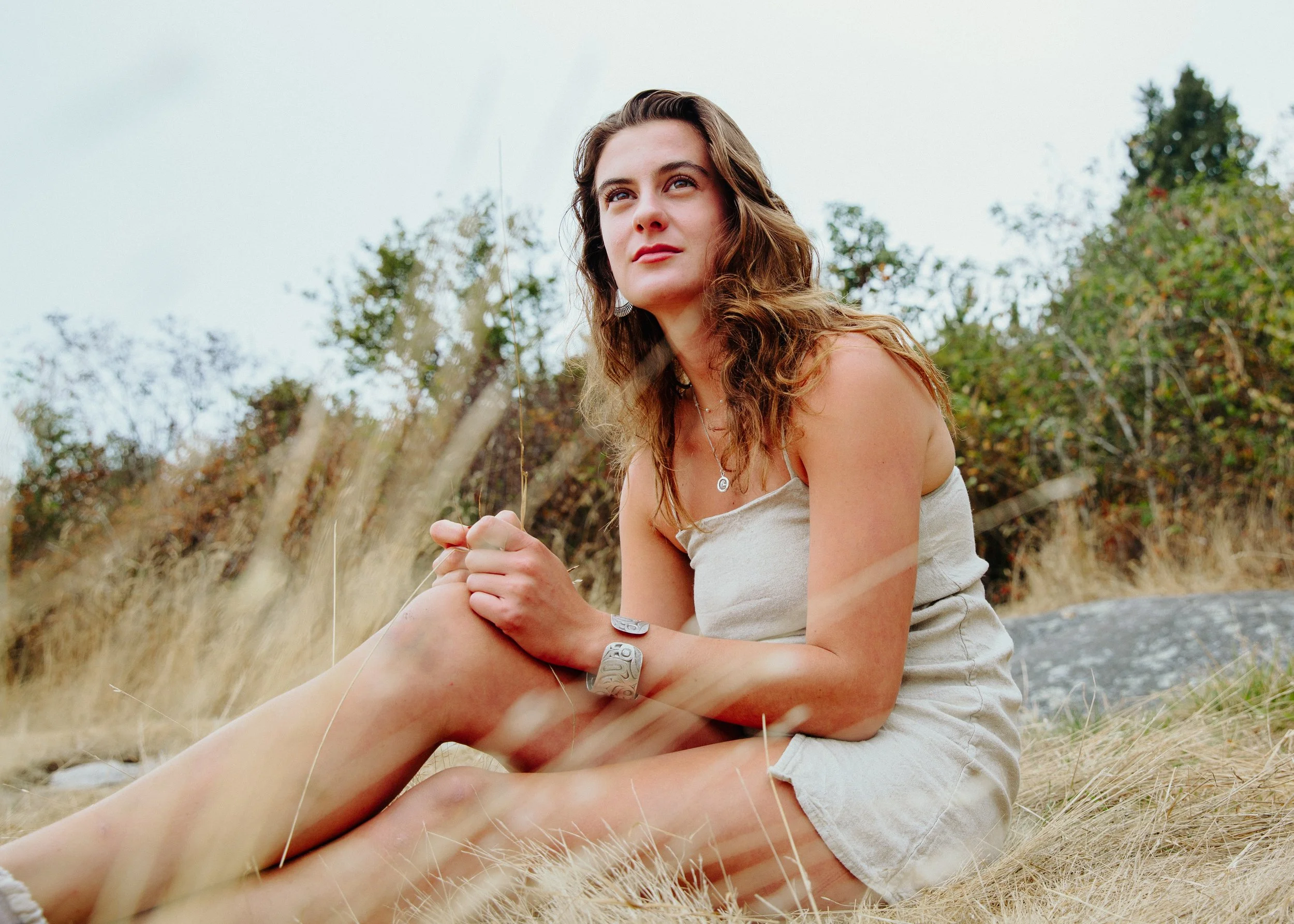 A young woman with wavy brown hair sitting outdoors in a natural setting with trees and tall grass, wearing a beige tank top and jewelry, gazing thoughtfully into the distance.