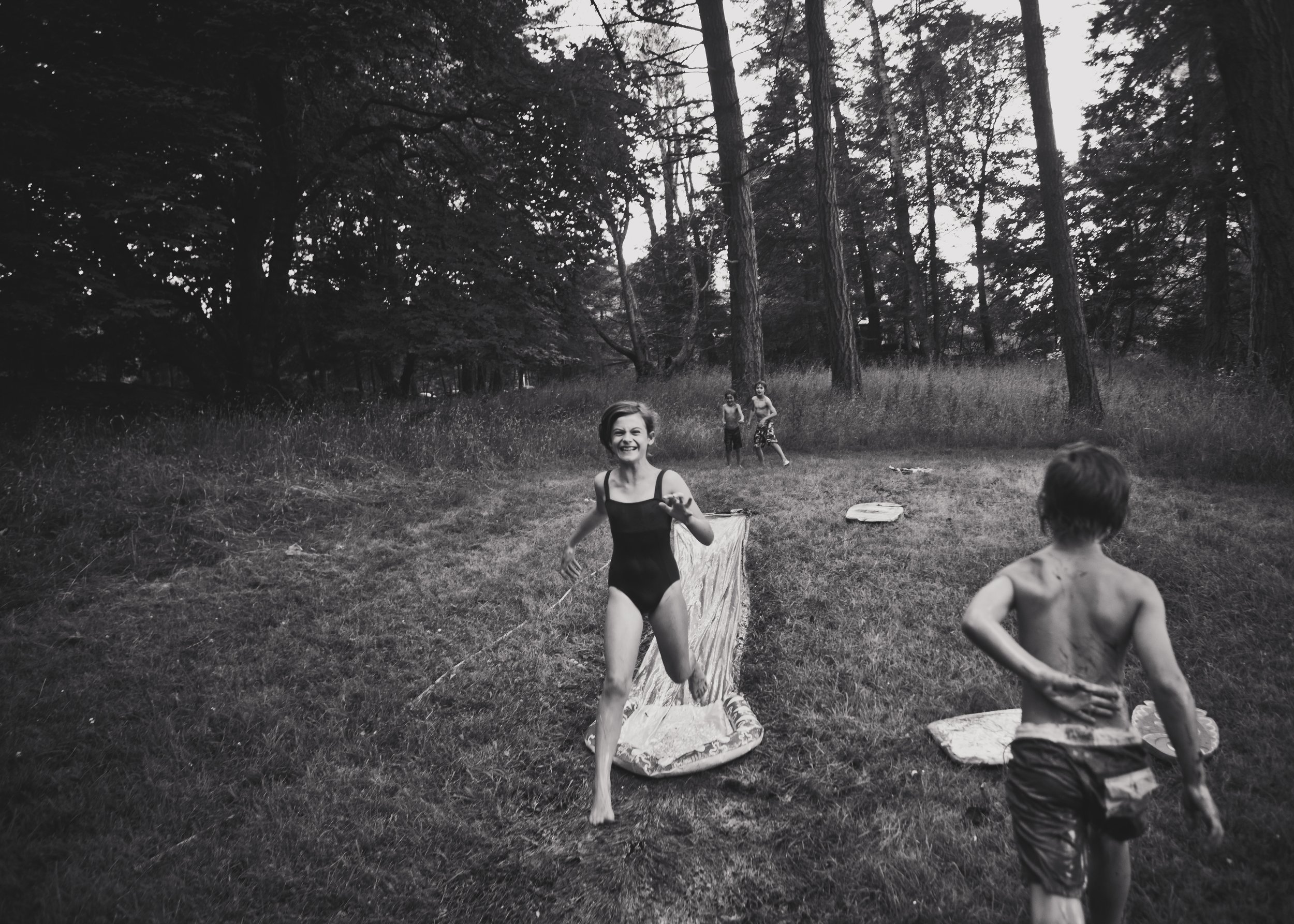 Black and white photo of children playing in a forest clearing, smiling and running with trees in the background.