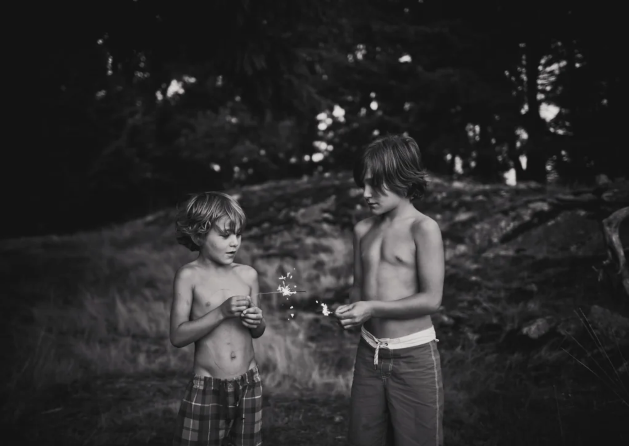 Two young boys standing outdoors at night holding lit sparklers. One boy is slightly taller and wearing striped shorts, the other is shorter and wearing plaid shorts. They are shirtless, with trees and dark foliage in the background.