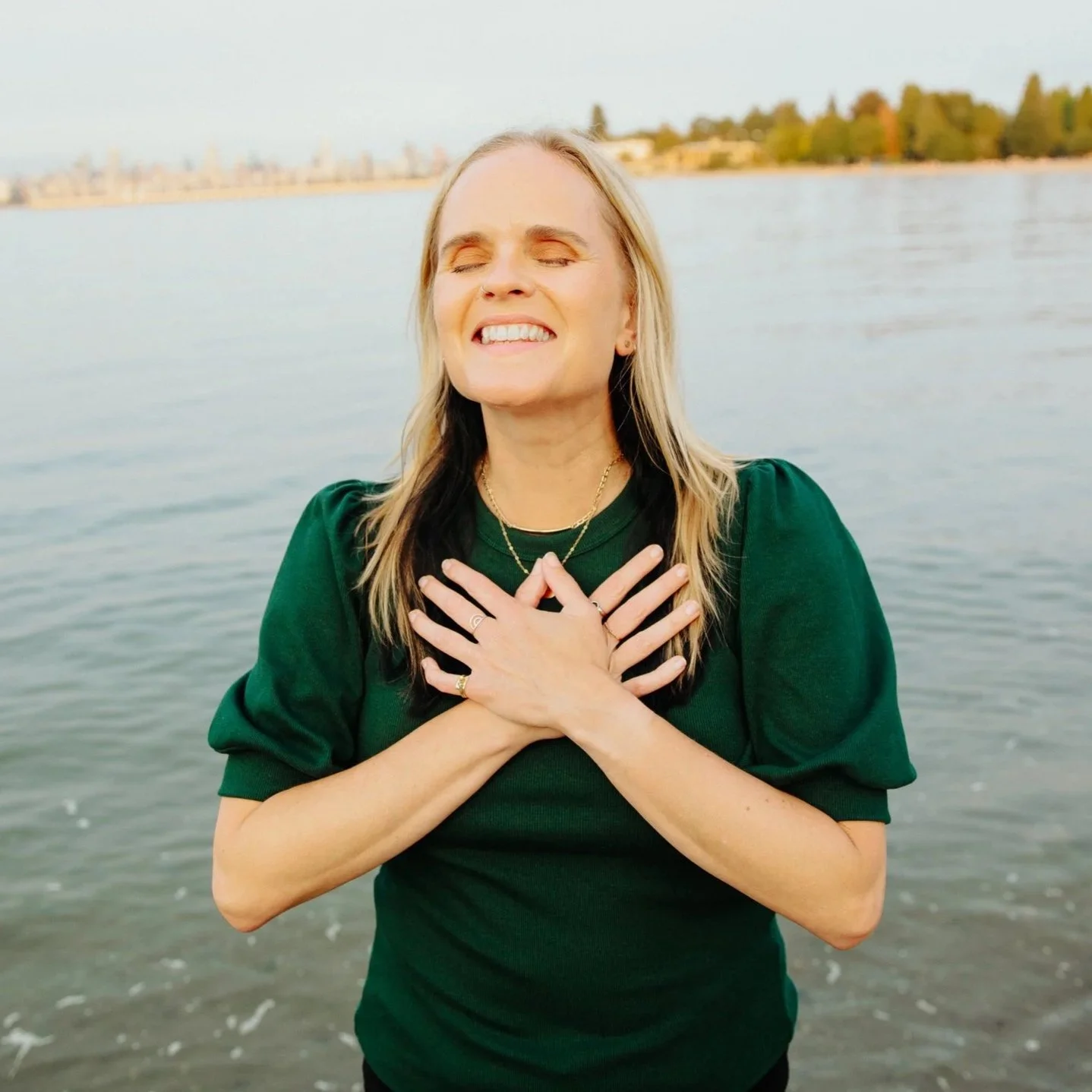 A woman with blonde hair in a green shirt, smiling with eyes closed, holding her hands over her chest, standing near a body of water with a cityscape in the background.