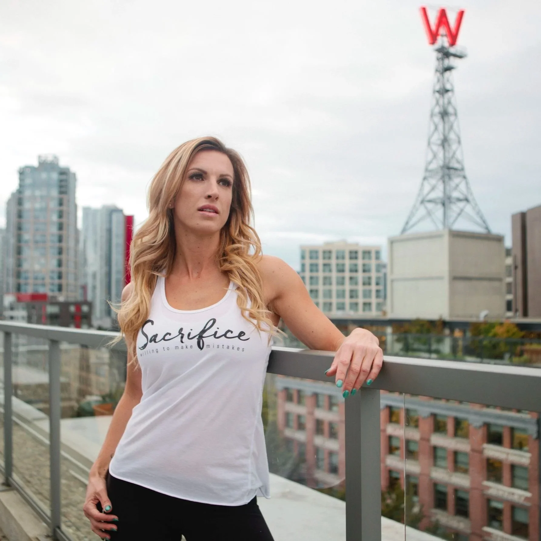A woman with long blond hair wearing a white tank top with the word 'Sacrifice' and words 'willing to make mistakes' writes on her nails while standing on a rooftop balcony in an urban setting with high-rise buildings and a large red 'W' sign in the 