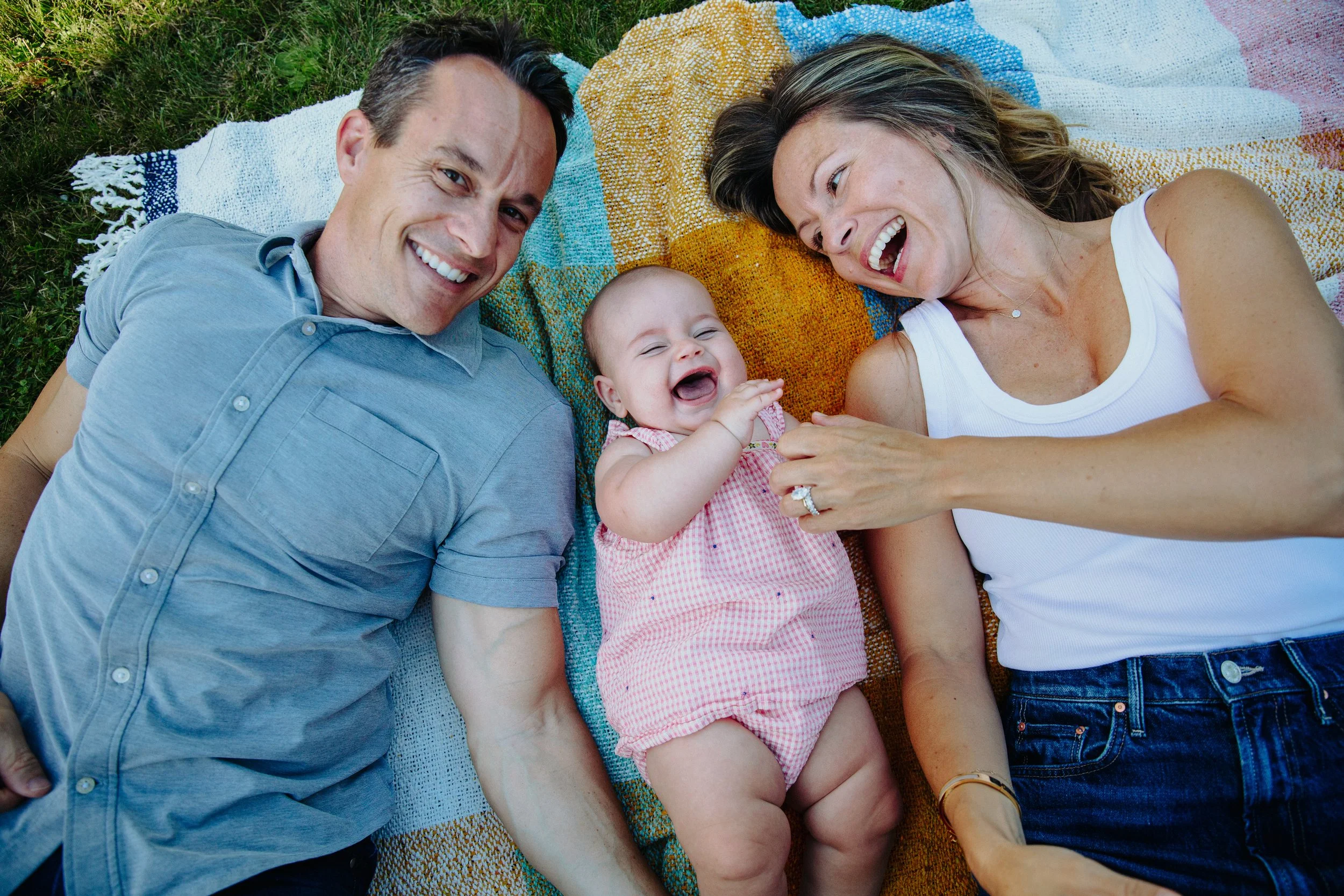 A happy family of three lying on a colorful blanket outdoors on grass, smiling and enjoying time together.