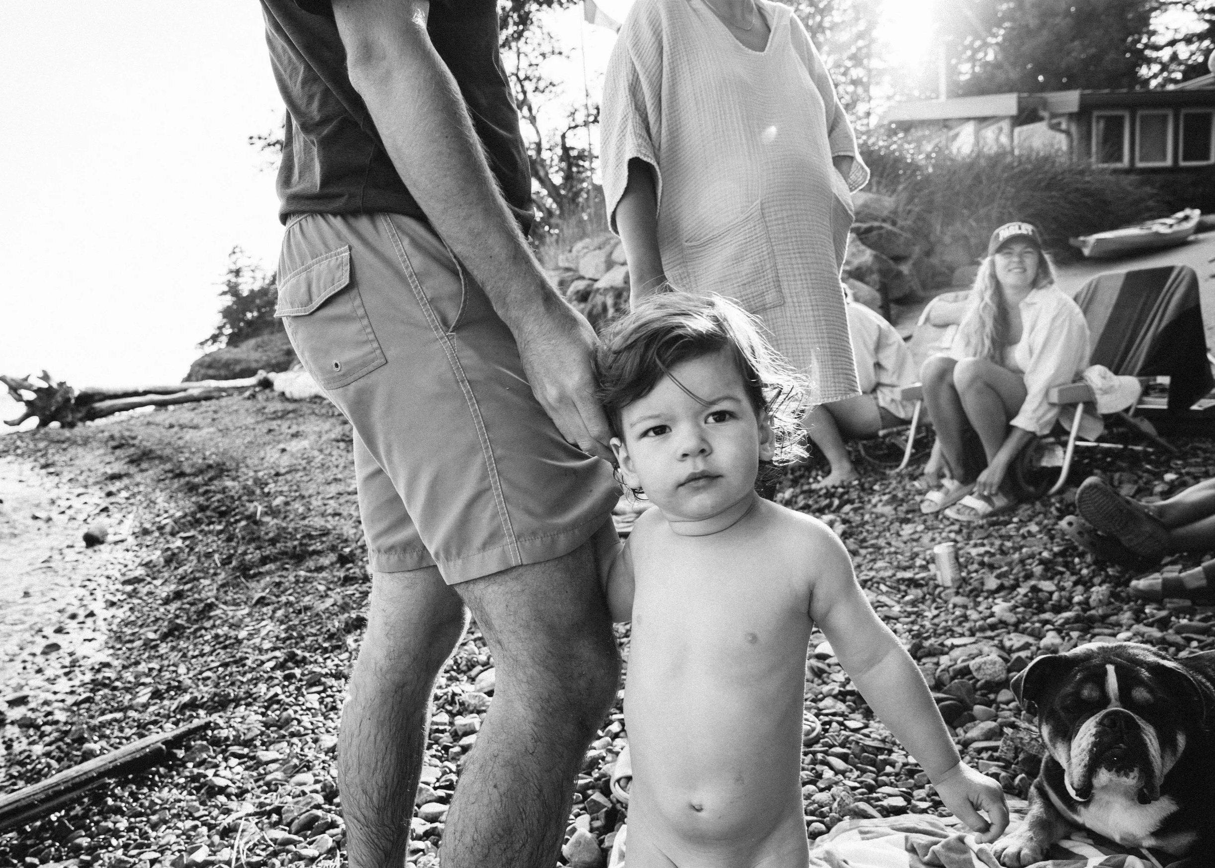 A black and white photo of a family outdoors at the beach. A young child standing shirtless, looking at the camera, is in the foreground. An adult, possibly a parent, holding his hand, is partially visible, with their torso in view. In the background
