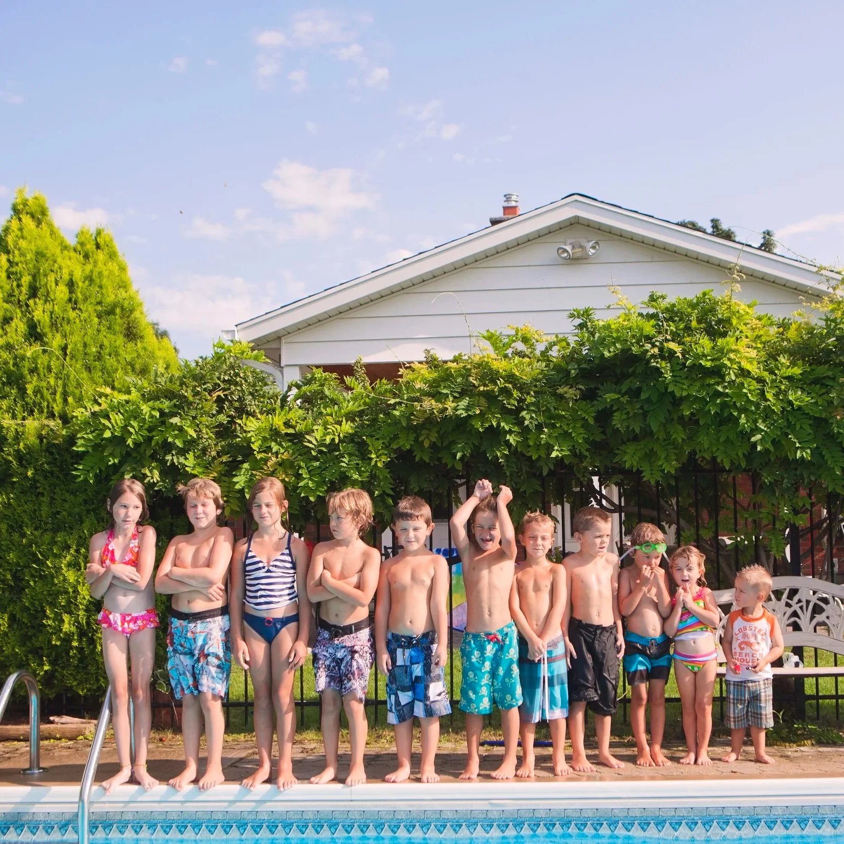Children wearing swimsuits standing by a swimming pool, with green shrubs and a white house in the background on a sunny day.