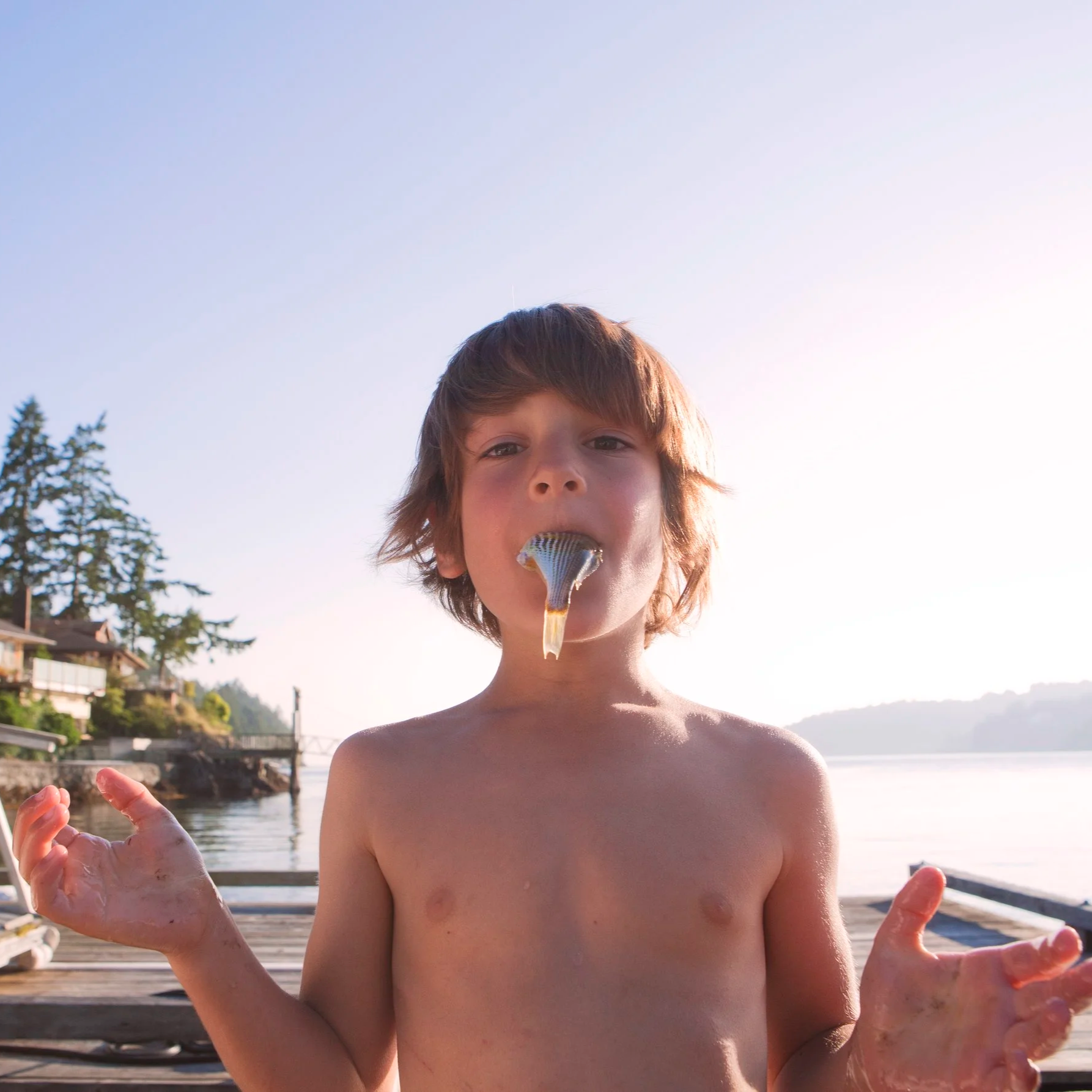 Young boy holding a fish in his mouth, standing outdoors near a body of water with trees and houses in the background during daytime.