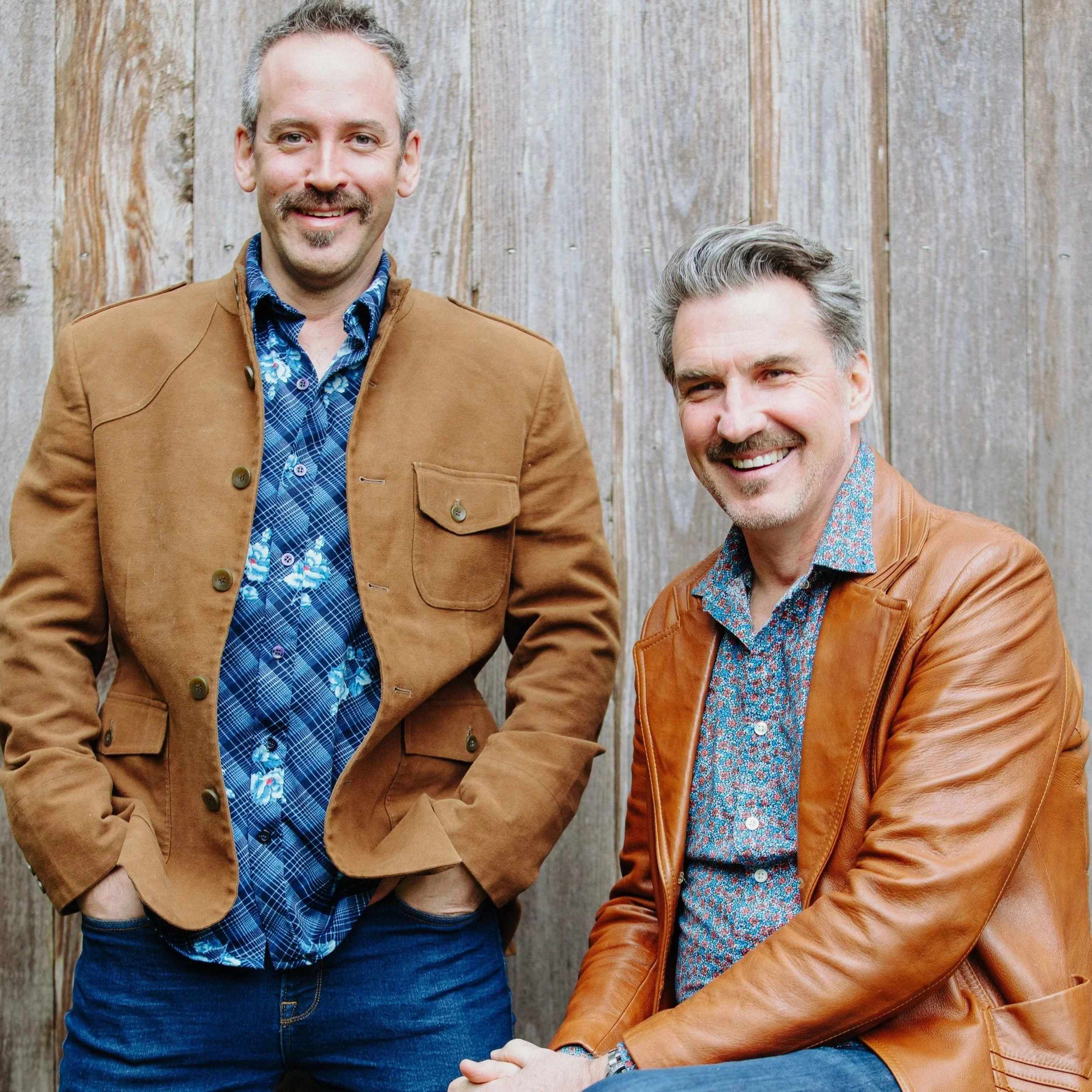 Close-up of two smiling men wearing casual clothing and jackets, standing in front of a wooden fence.