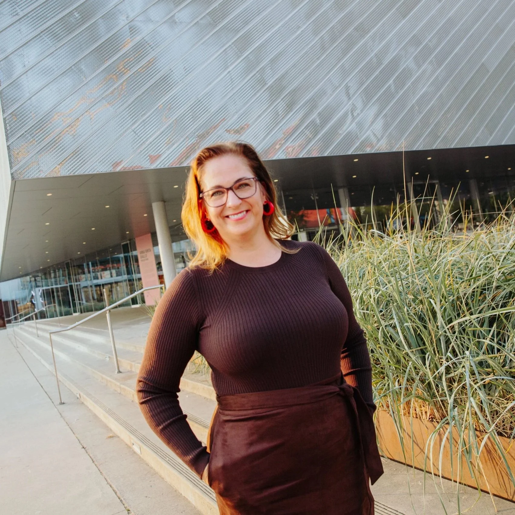 Woman in a black top and dark skirt standing outdoors in front of a modern building with glass windows and metallic panels, with plants in the foreground.