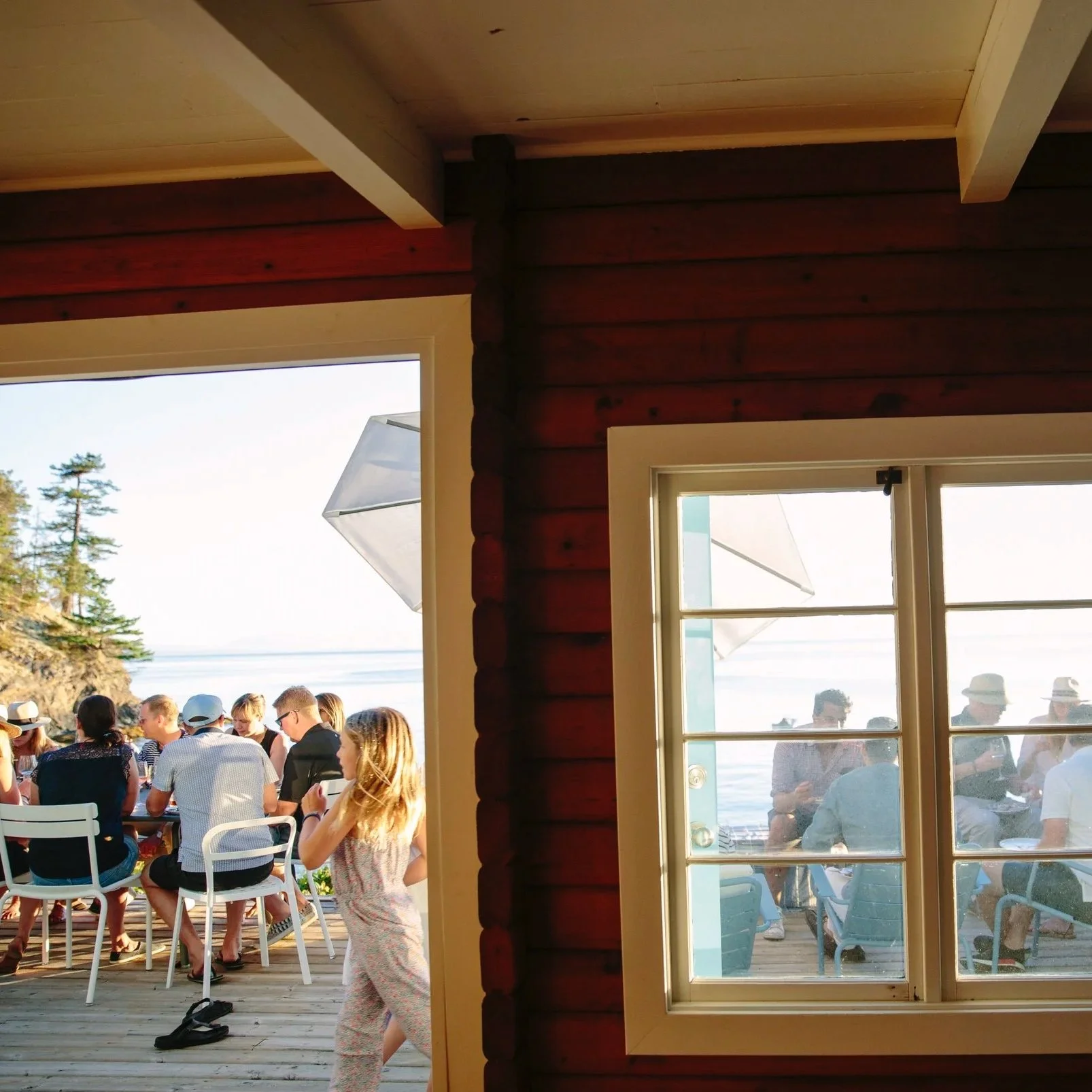 People gathered outdoors on a deck near the water, seen through a window and door of a wooden cabin.