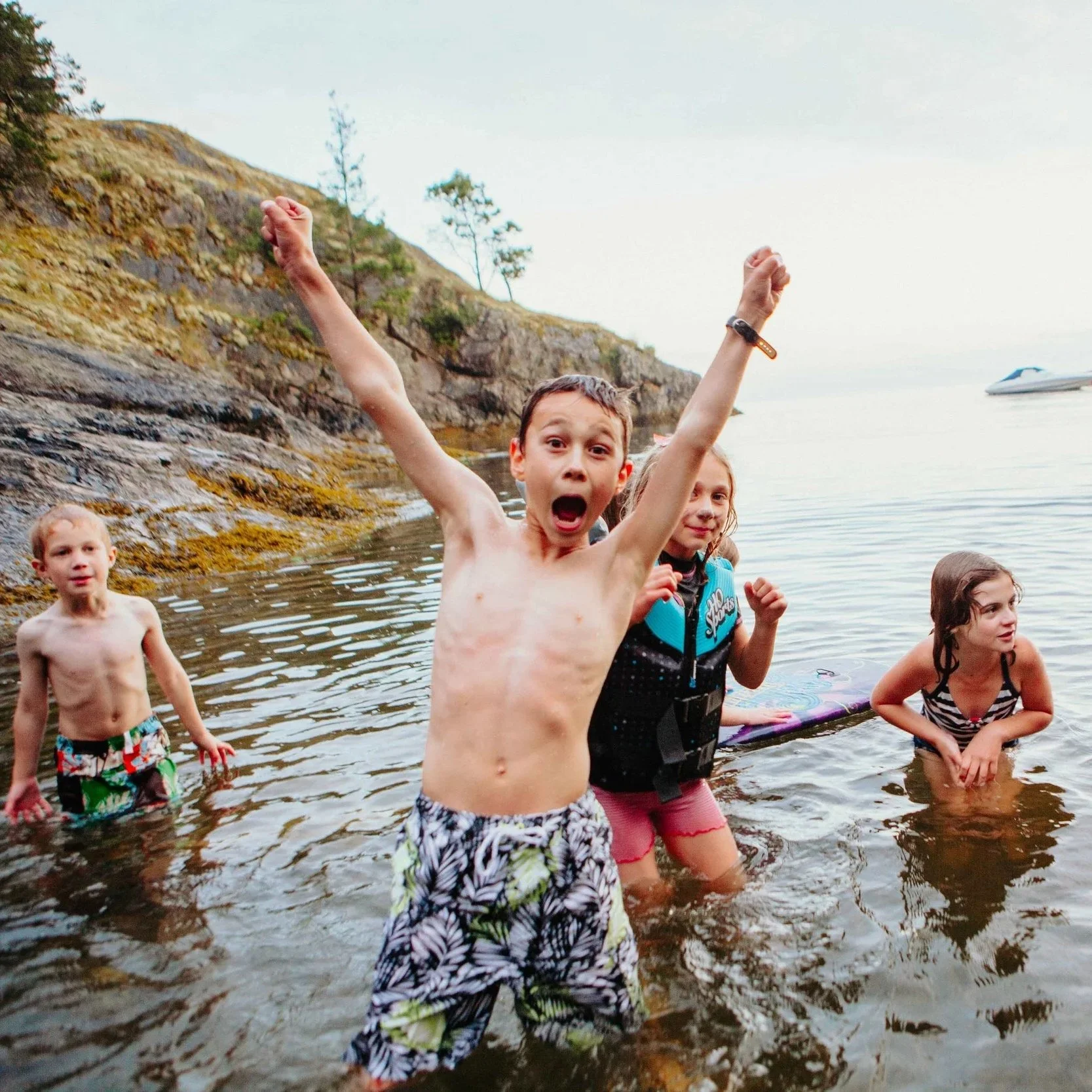 Group of children playing and splashing in a shallow rocky beach with hills and trees in the background.
