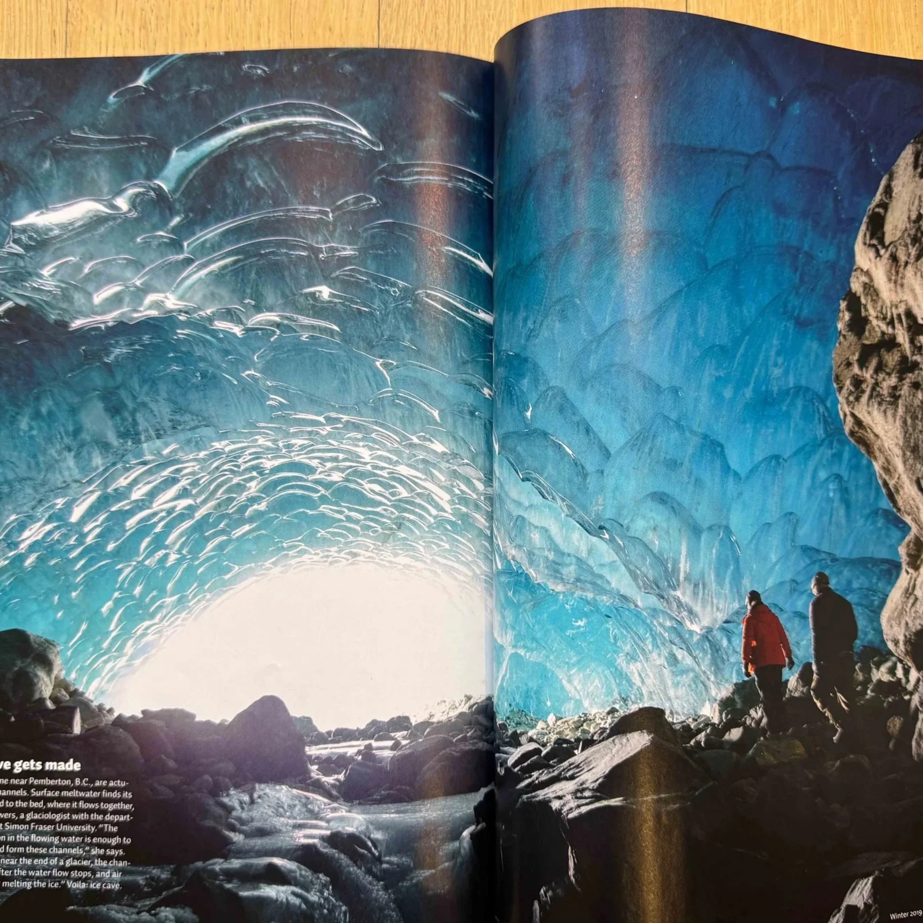 A large ice cave with blue and white ice formations on the ceiling and walls, and two people in winter clothing standing on rocky ground inside the cave.