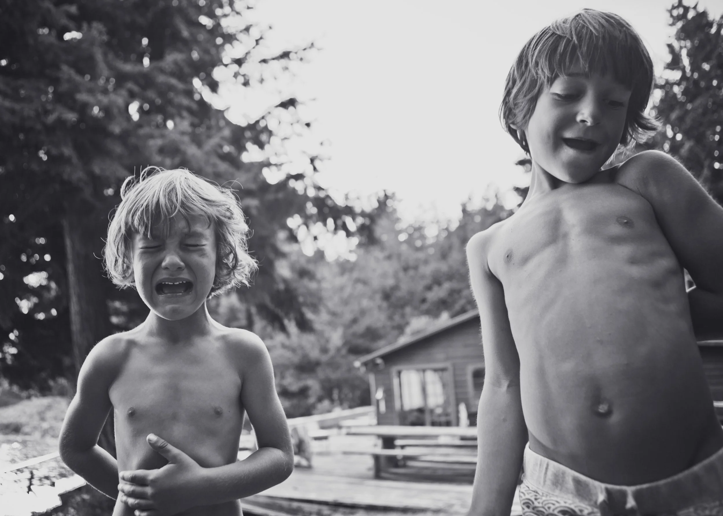 Two young boys with wet hair and no shirts, one crying and the other looking surprised or scared, outdoors near a wooden structure and trees.