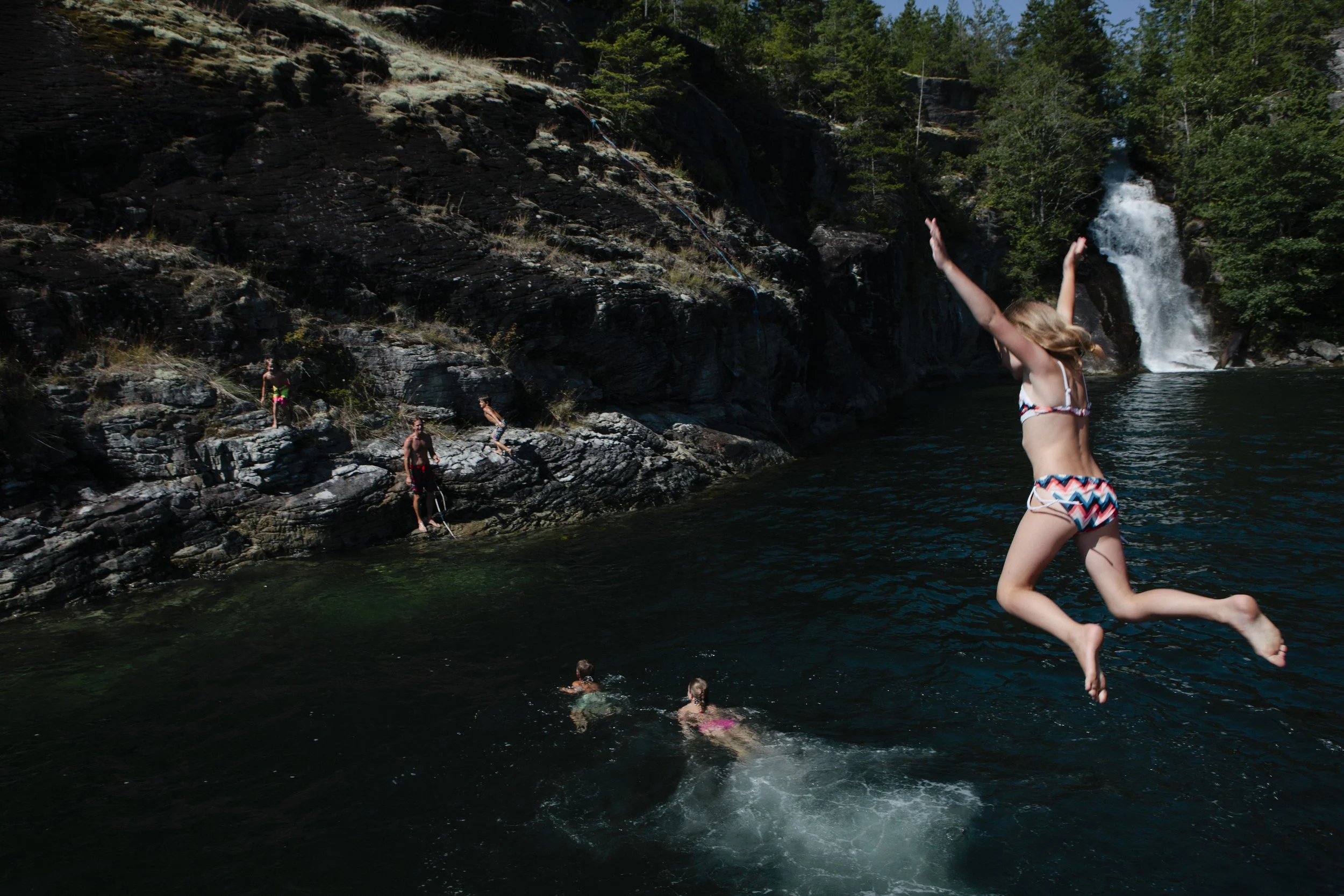 Children swimming and playing in a natural river near a waterfall surrounded by trees and rocky cliffs.