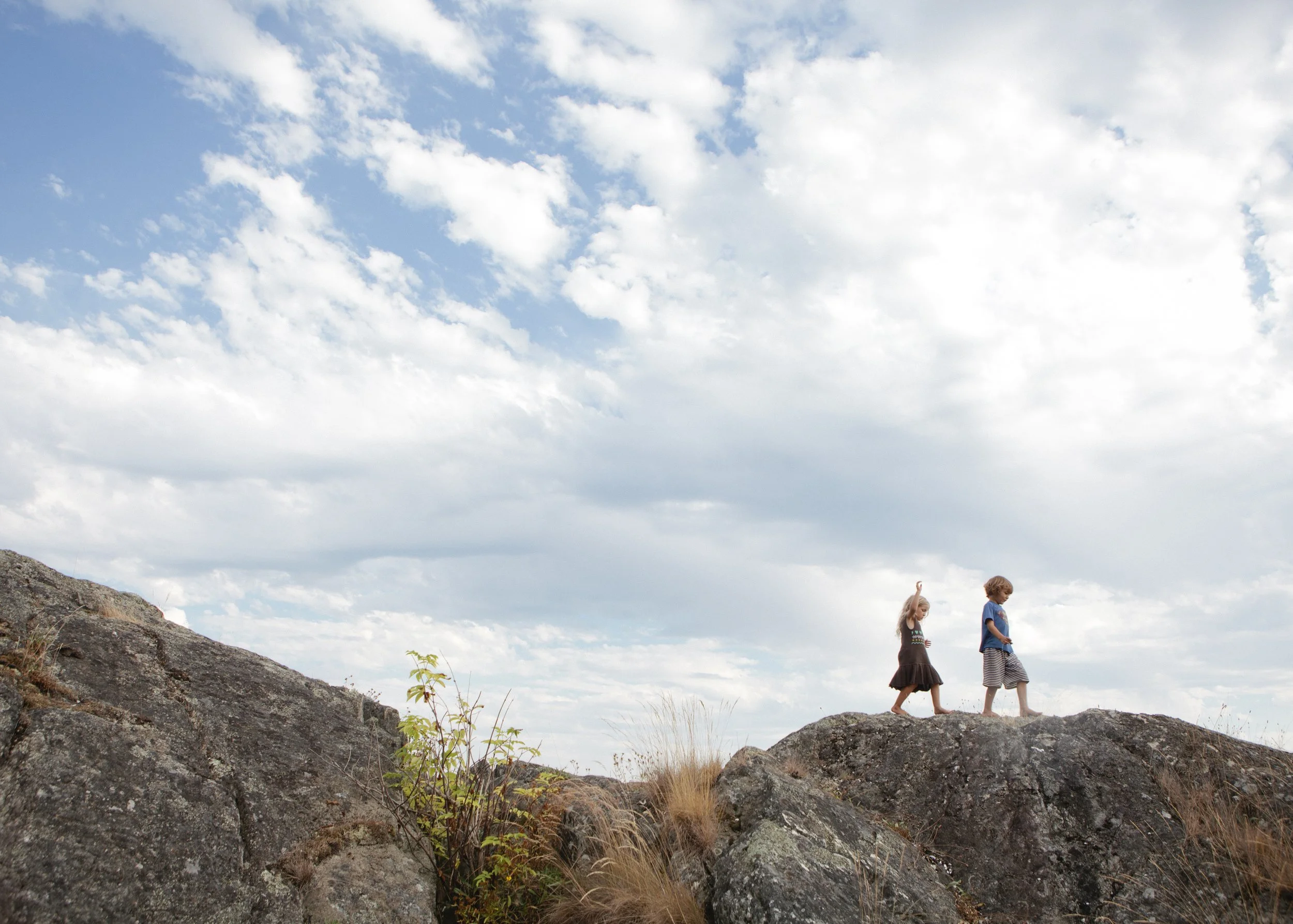 Two children walking barefoot on rocks outdoors under a partly cloudy sky.
