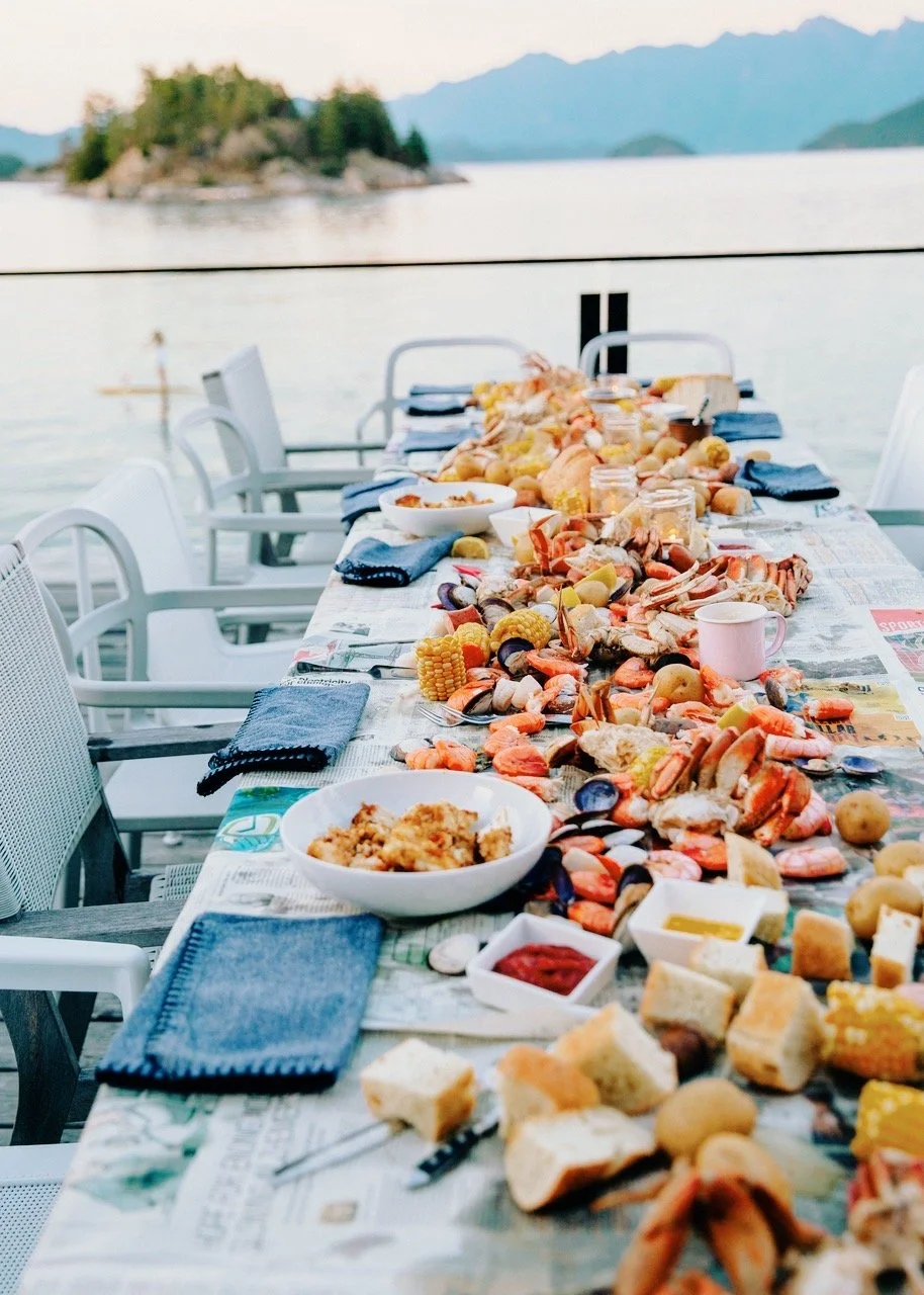 A long outdoor table set for a seafood feast overlooking a large body of water and mountains in the background, with blue napkins and plates.