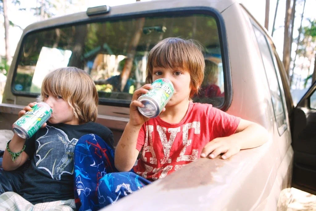 Two young boys sitting on the hood of a truck, drinking from soda cans, outdoors with trees in the background