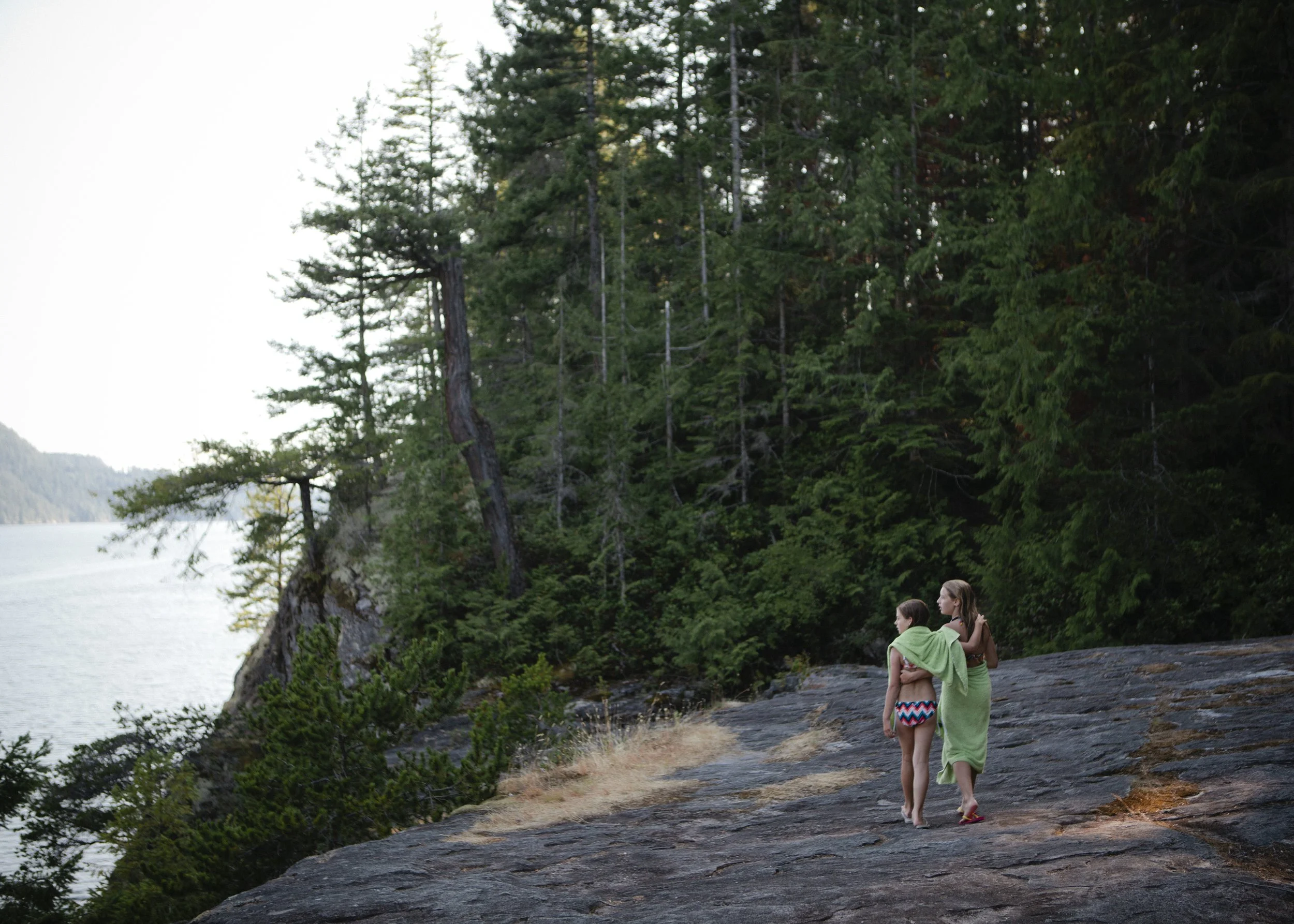 Two young girls wrapped in a green towel standing on a rocky shoreline near a dense forest and a body of water, with trees and hills in the background.