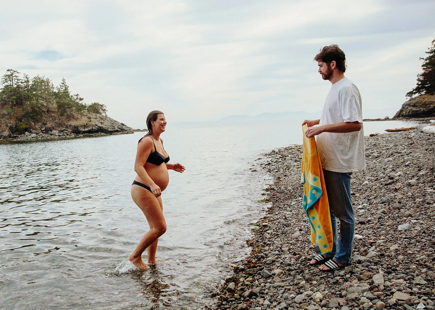 A pregnant woman in a black bikini wading into the water at the beach, smiling, as a man in a white t-shirt and jeans holding a towel looks on.