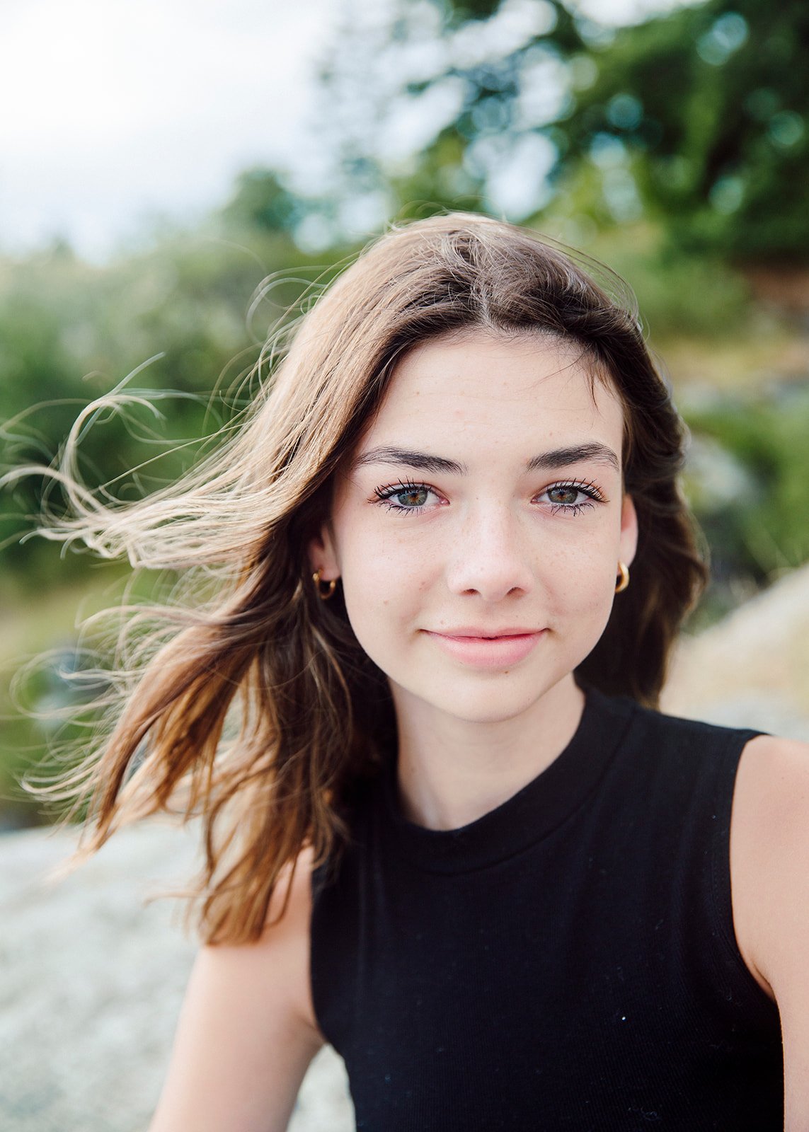 A young woman with long, wavy brown hair and green eyes smiling outdoors with a blurred green background.
