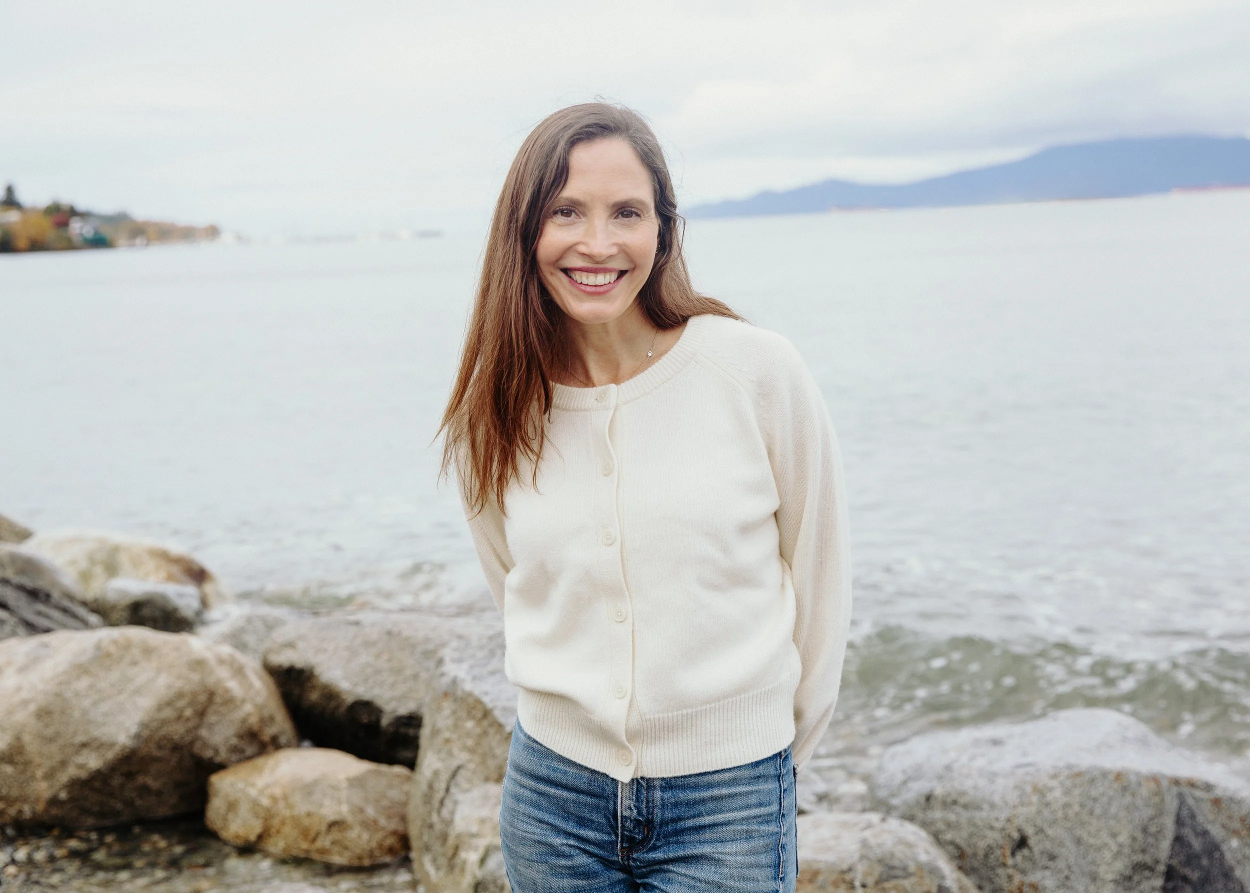A woman with long brown hair smiling at the camera while standing by a body of water with rocks and mountains in the background.