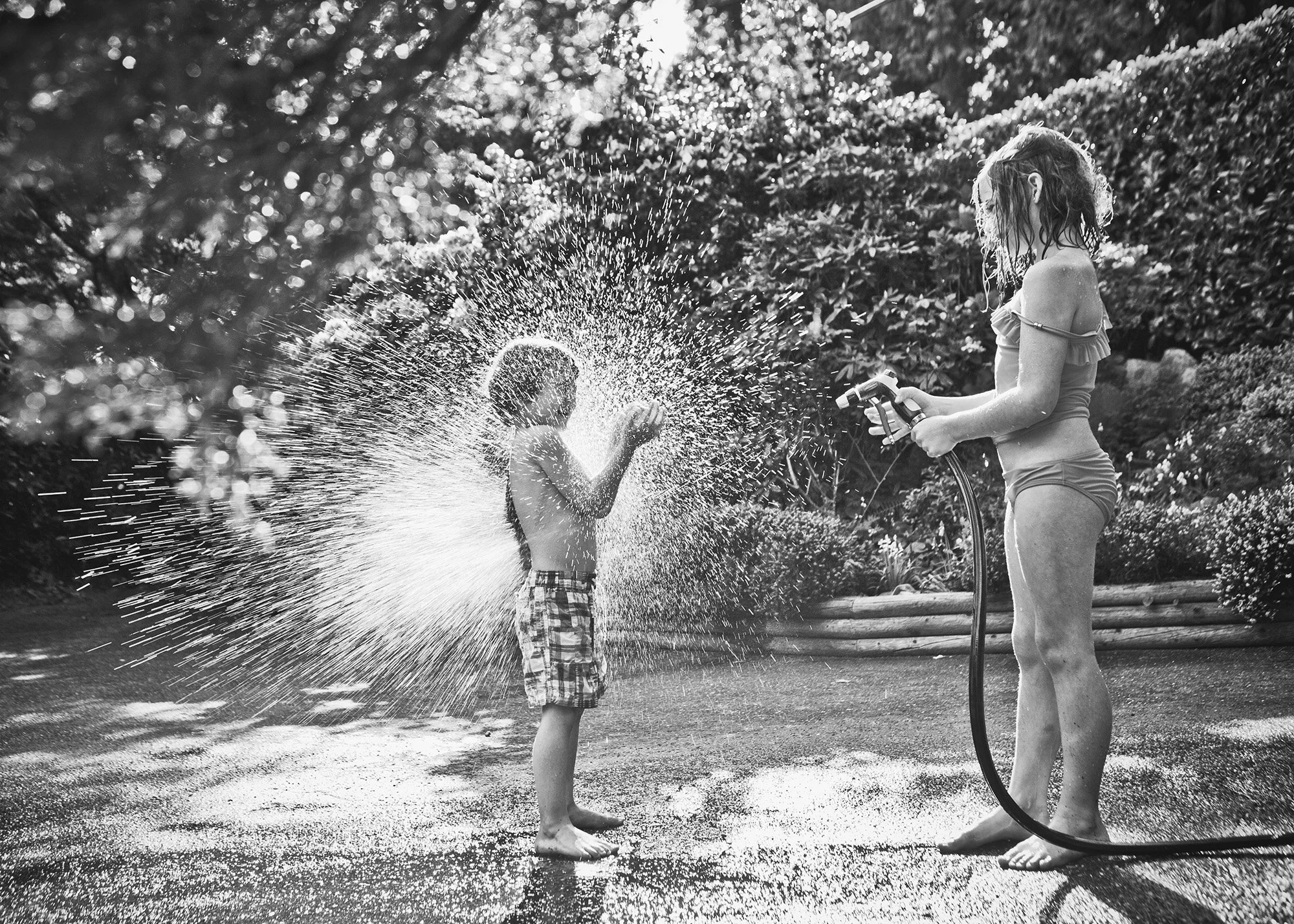 A girl and a boy playing with a garden hose, splashing water on each other in a backyard with lush greenery, captured in black and white.