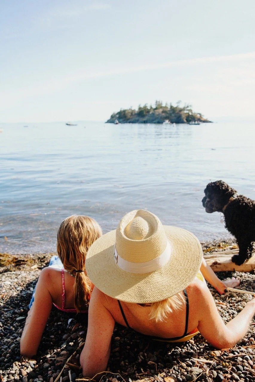 A woman in a sunhat relaxing on a pebbled beach next to a young girl and a black dog, with water and an island in the background.