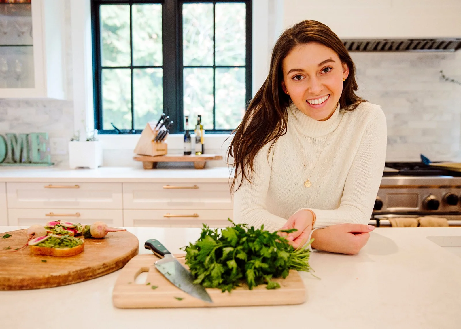A young woman with long brown hair smiling in a bright kitchen, preparing fresh herbs on a kitchen island with a knife and a wooden cutting board, with a window showing green trees in the background.