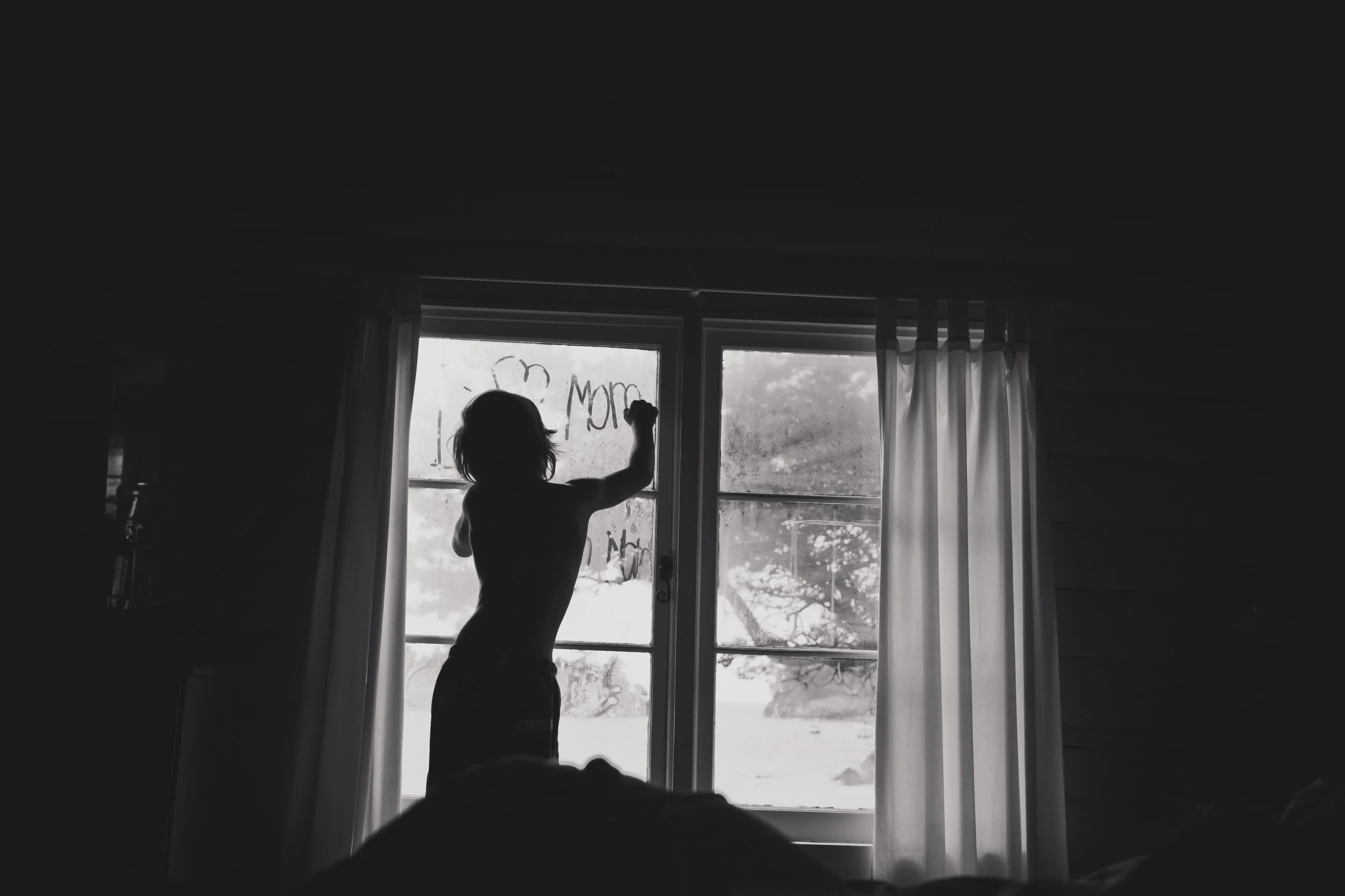 Child drawing on a window with rain outside, with curtains partially open.
