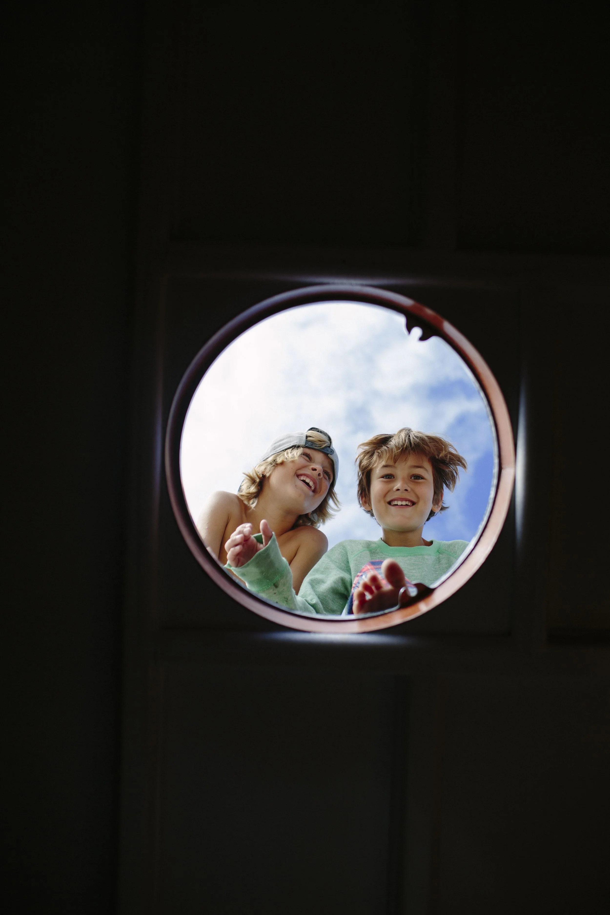 Two children looking down into a circular opening, with blue sky and clouds visible in the background, smiling and appearing happy.